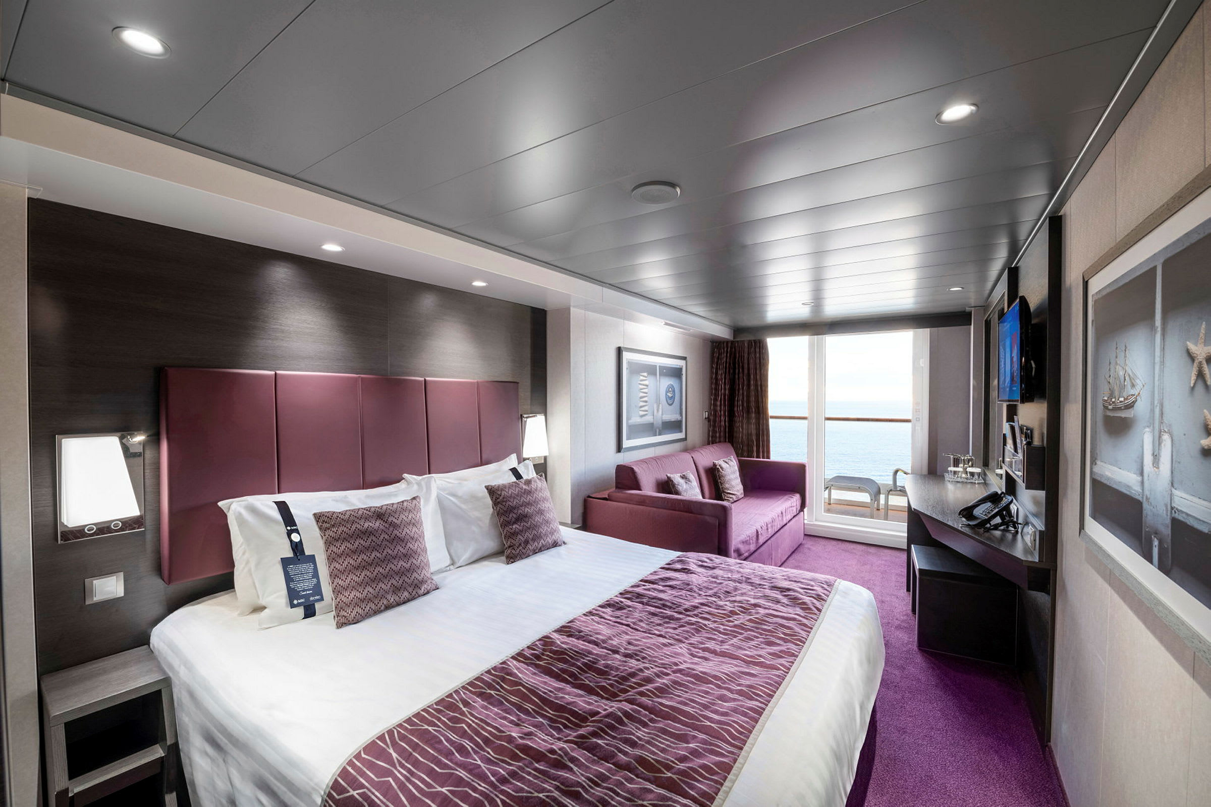 Ett hotellrum med en stor säng, en soffa, skrivbord och utsikt genom glasdörrar mot en balkong med stolar. (AI-genererad text)