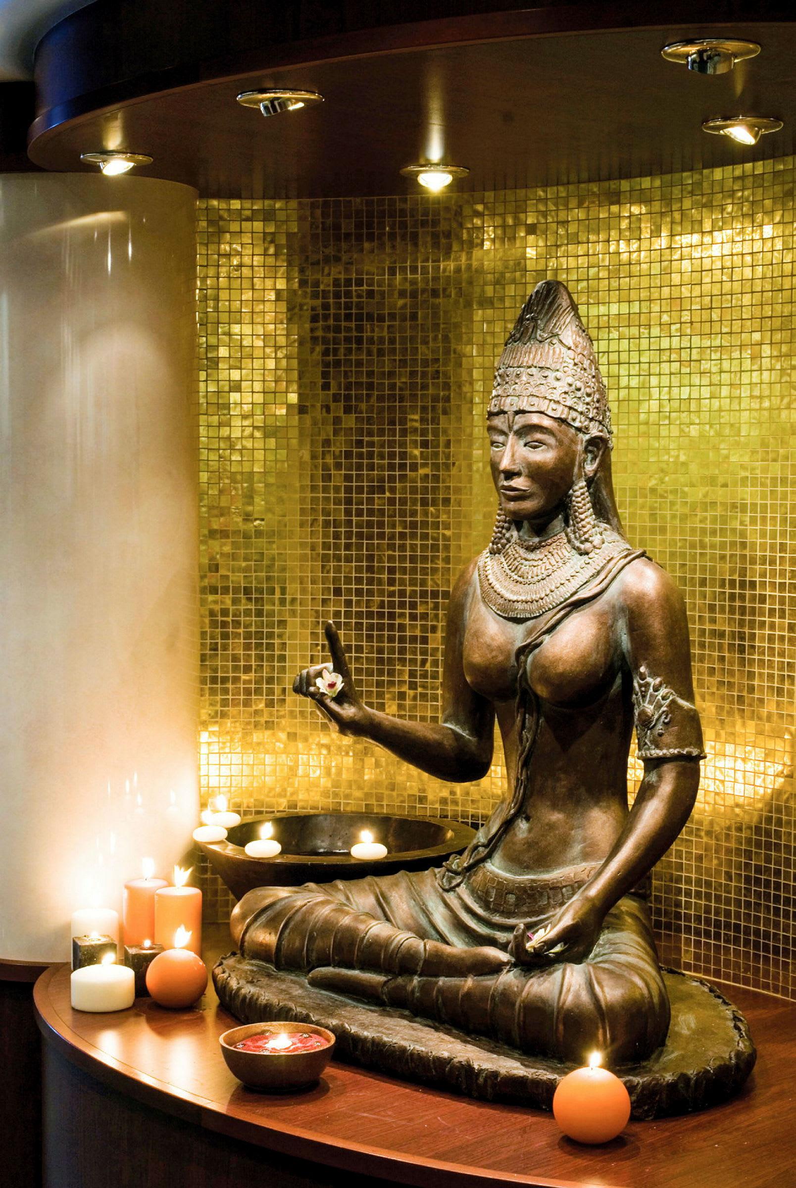 Staty av en buddhistisk figur framför en gyllene mosaikvägg omgiven av tända ljus. (AI-genererad text)