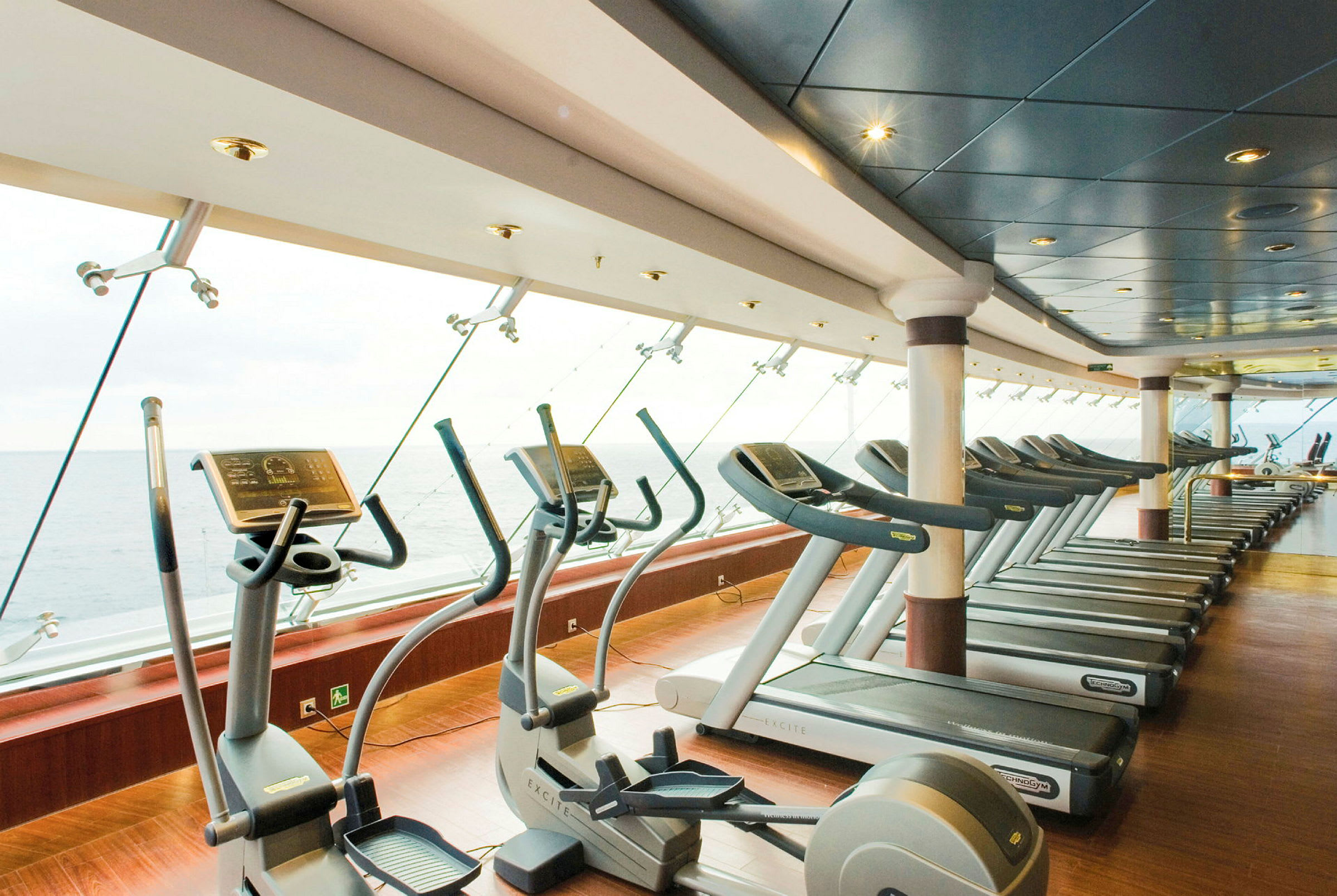 Ett gym med löpband och träningsmaskiner framför stora fönster med utsikt över havet. (AI-genererad text)