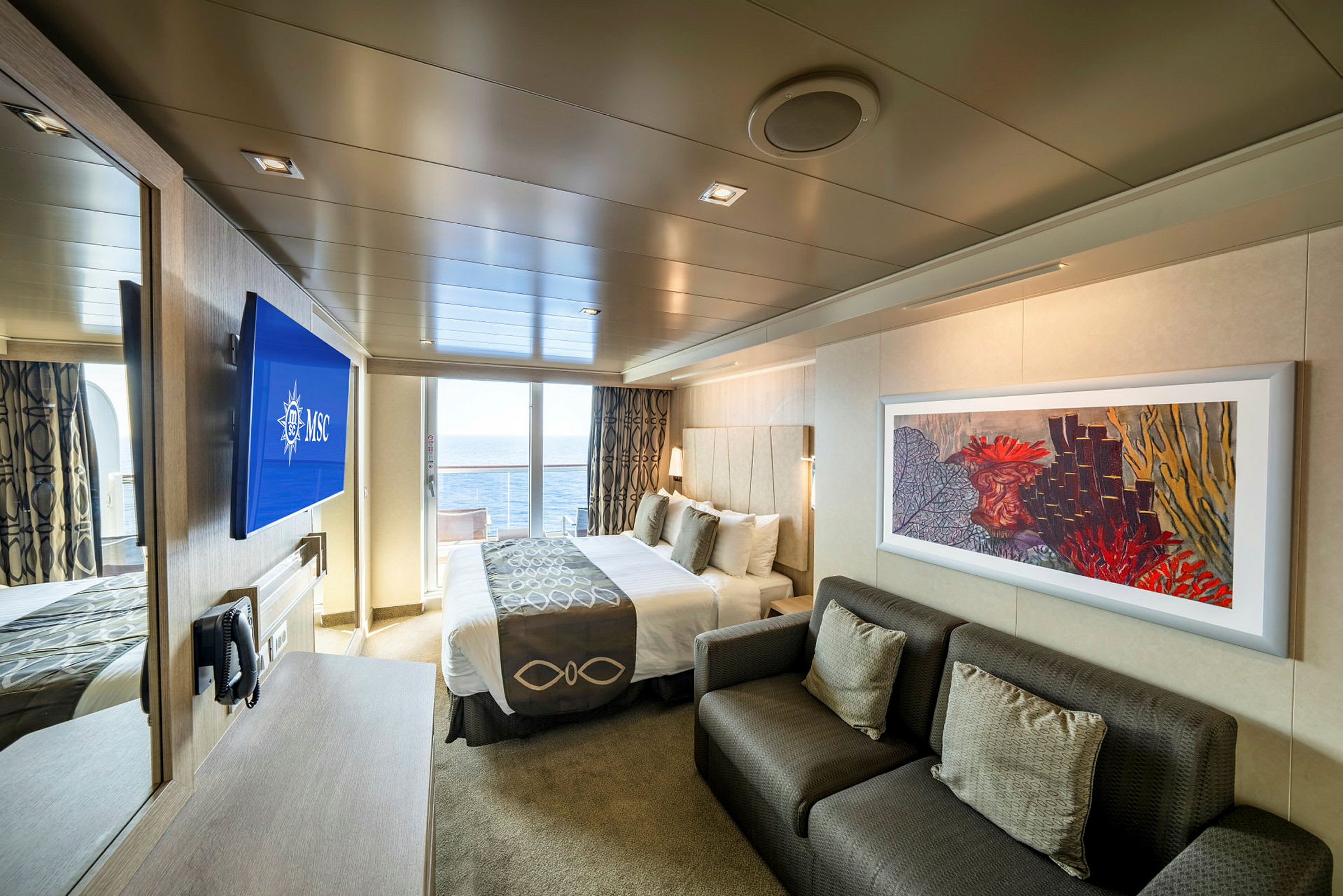 En modern hotellsvit med havsutsikt genom glasdörrar, inklusive en dubbel säng, en soffa och en vägg-tv. (AI-genererad text)