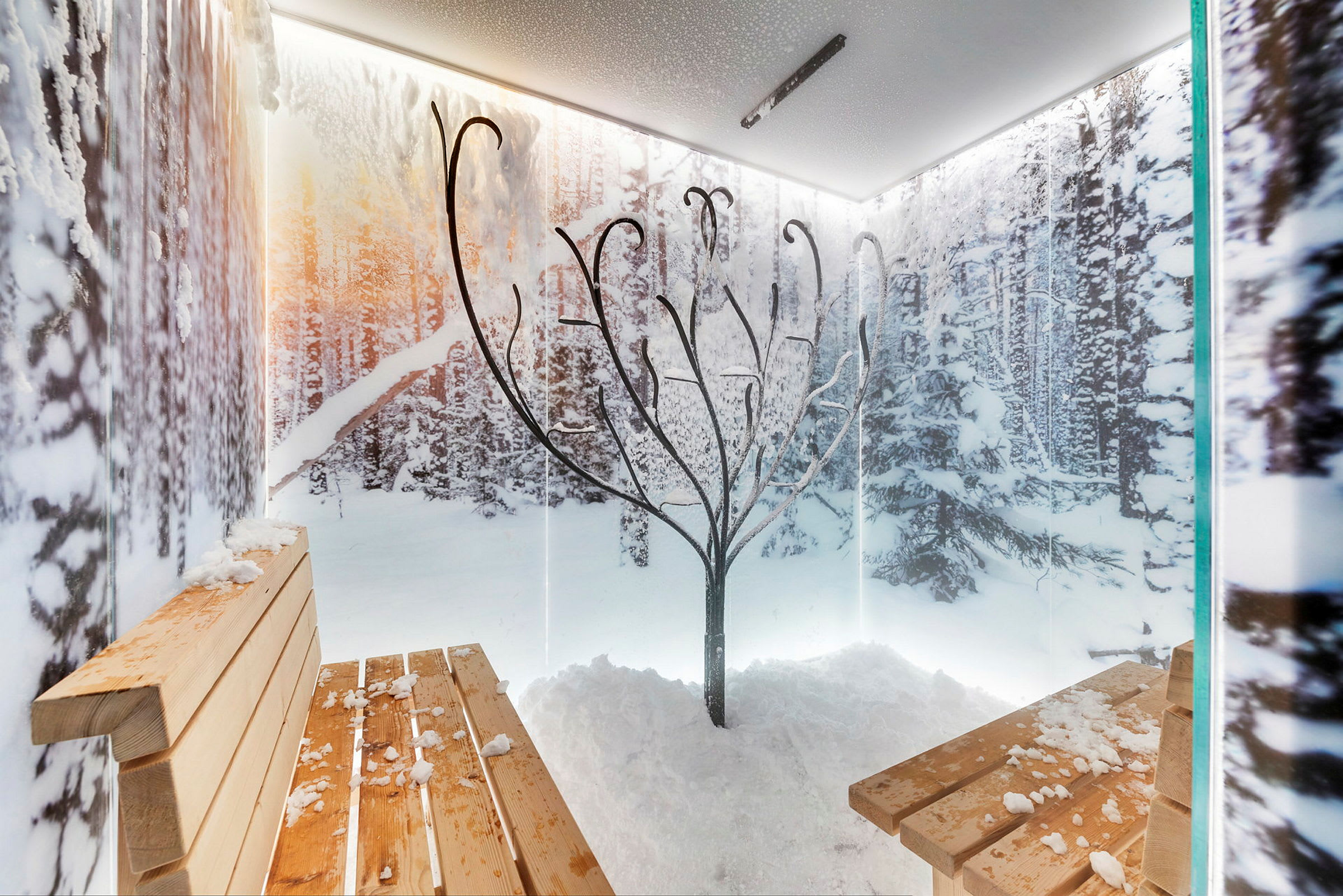 En interiör med bänkar av trä, en central metallkonst med grenliknande strukturer och snötäckta detaljer, omgiven av väggar med mönster av vinterlandskap. (AI-genererad text)