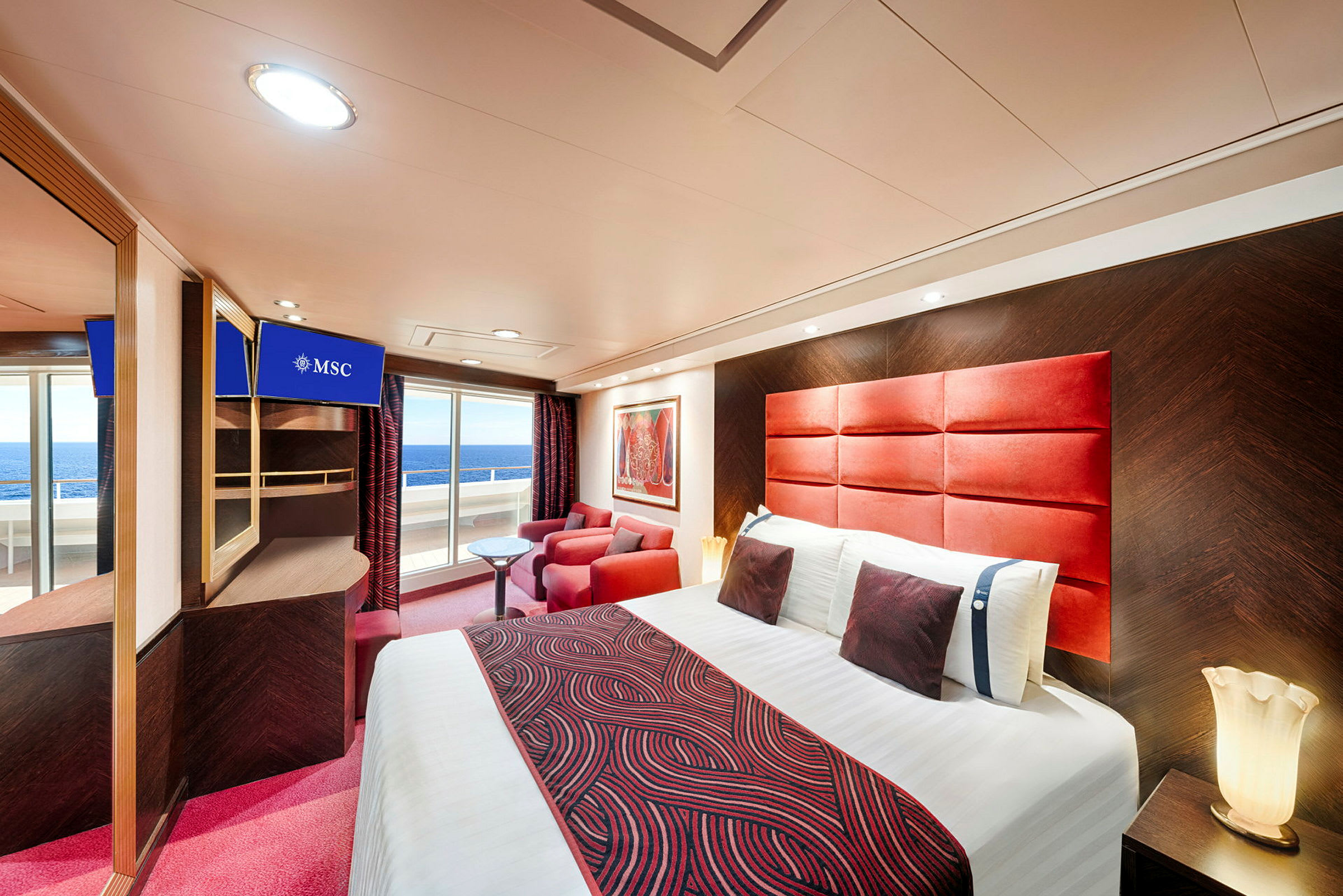 Ett modernt hotellrum med en dubbelsäng, rödmönstrade textilier, röda fåtöljer, ett litet bord och en balkong med utsikt över havet. (AI-genererad text)