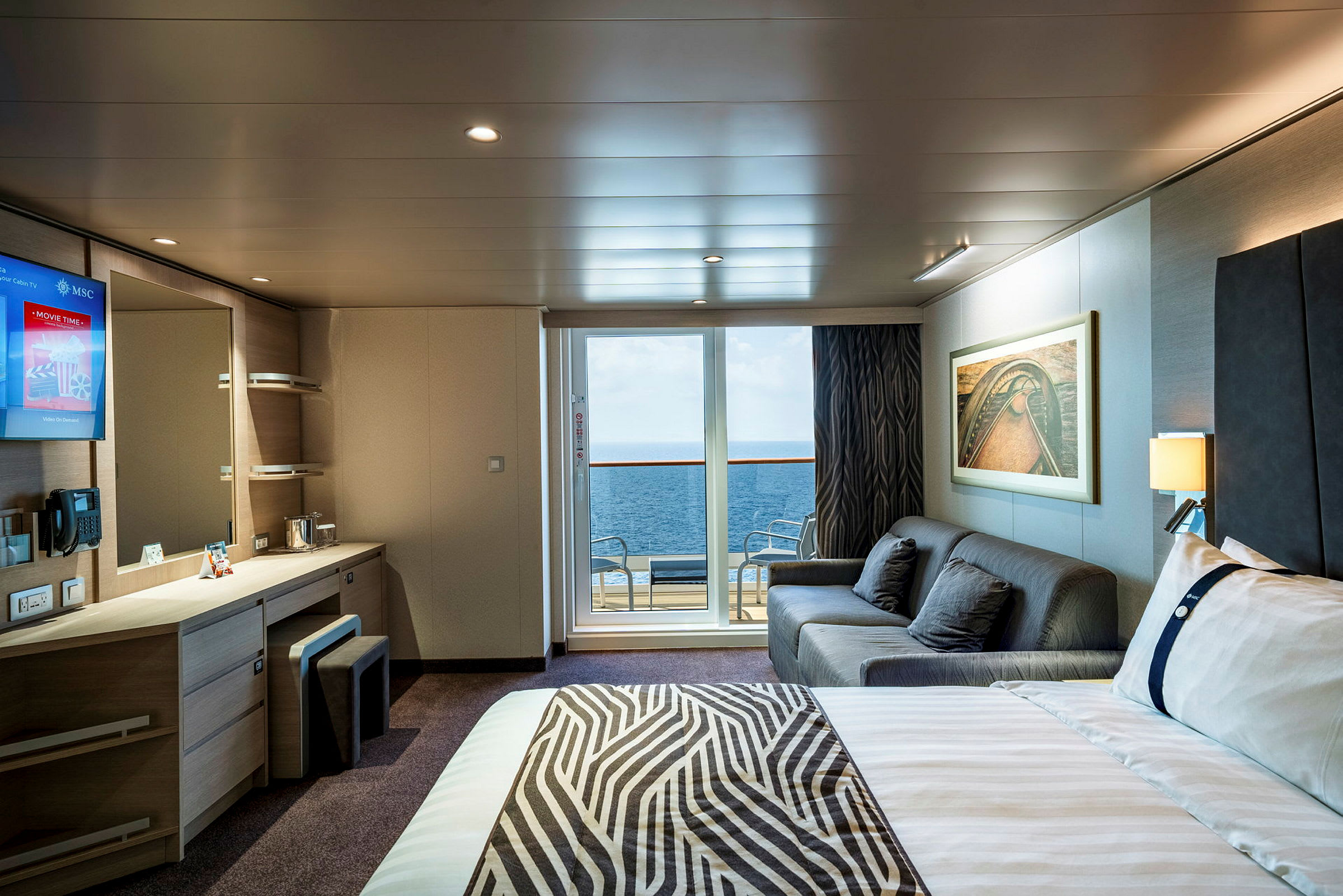 En interiör av ett hotellrum med säng, soffa, skrivbord och en balkong som har utsikt över havet. (AI-genererad text)