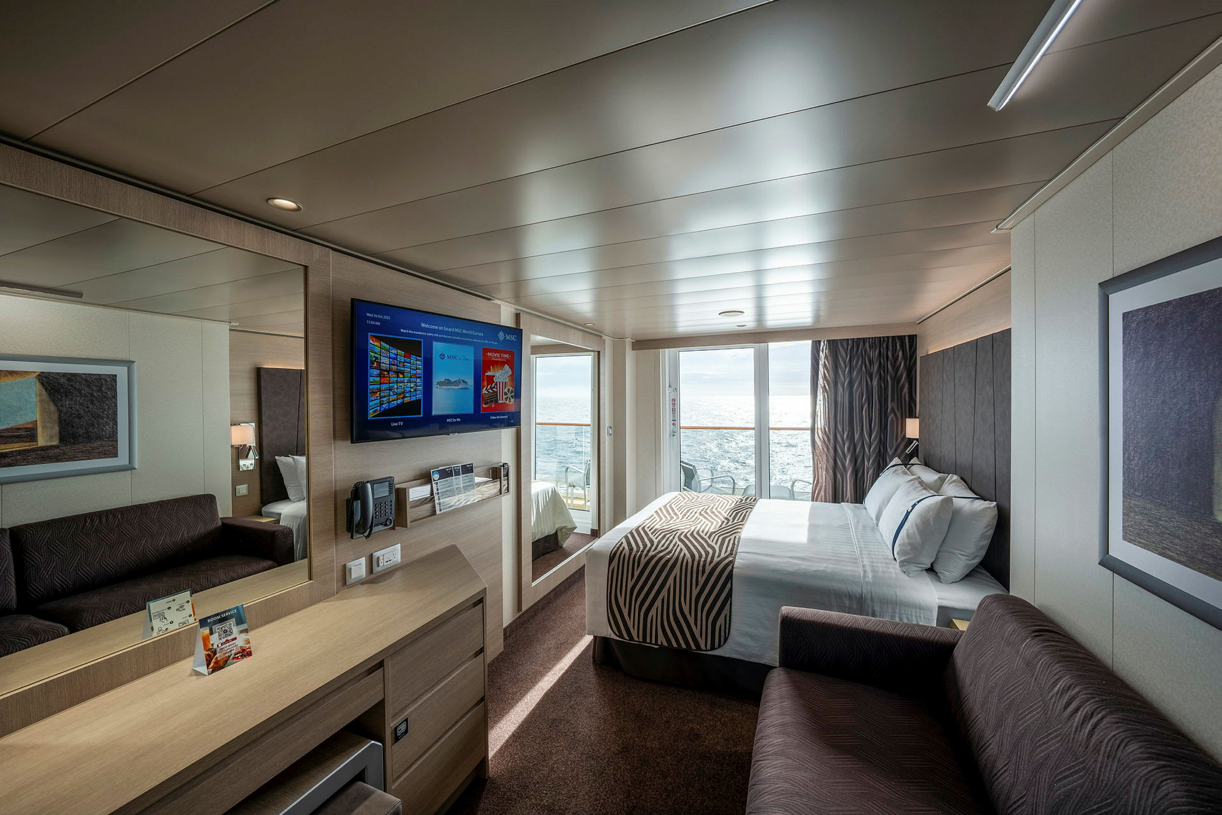 Modernt inrett hotellrum med dubbelsäng, soffa, skrivbord och väggmonterad TV. Stor glasdörr leder till balkong med havsutsikt. (AI-genererad text)
