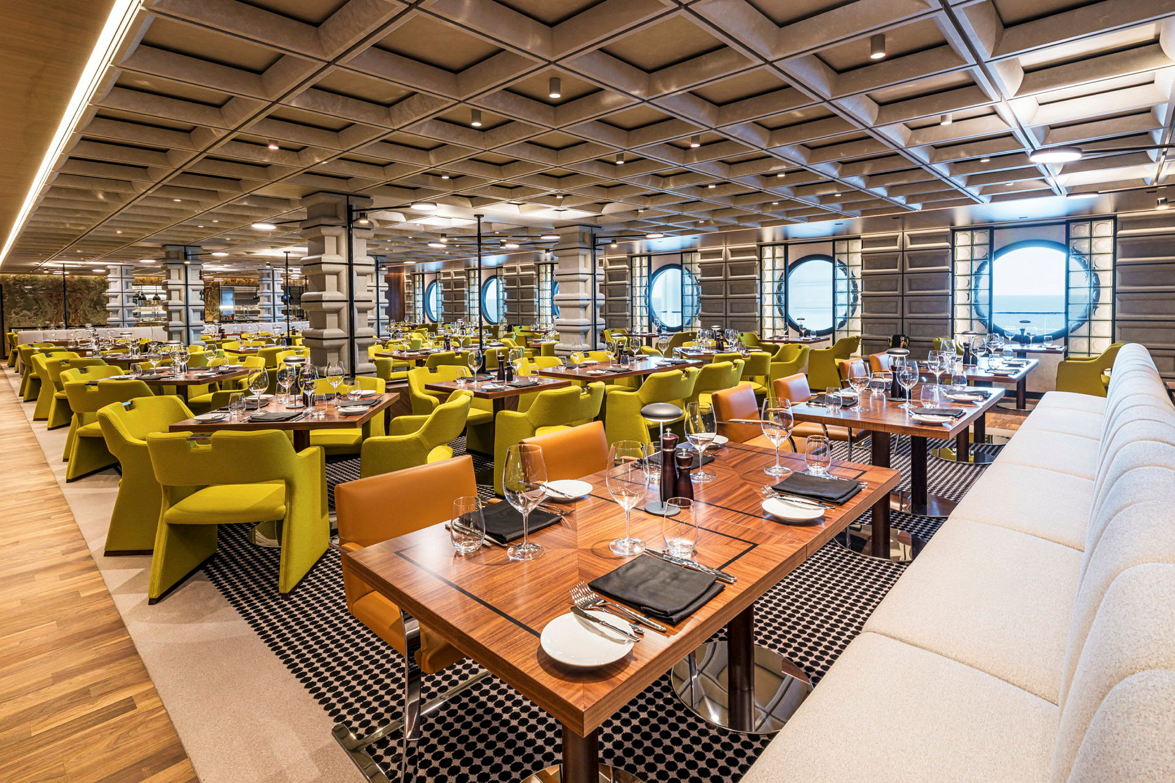 Interiören av en modern restaurang med många dukade bord, gröna stolar och en välvd vägg med runda fönster. (AI-genererad text)