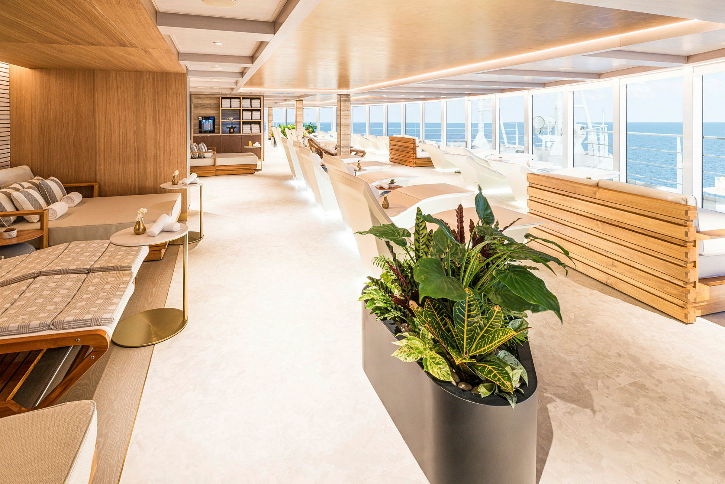 Ljus och modern lounge med liggstolar, soffor och stora fönster med utsikt över havet. Växter i förgrunden. (AI-genererad text)