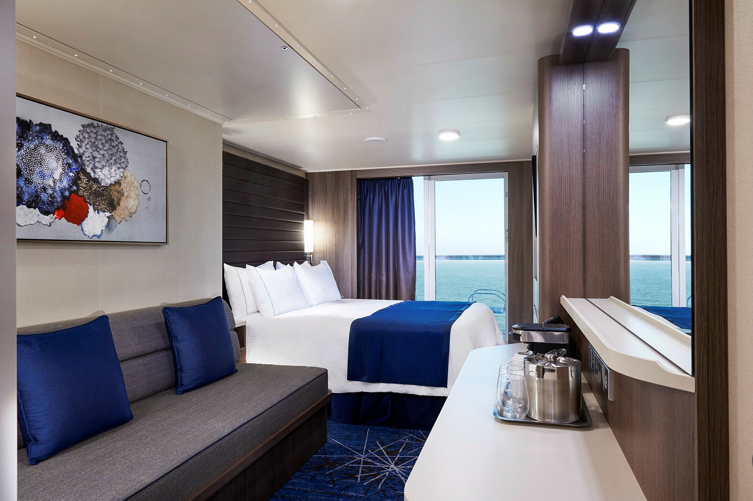 Ett hotellrum med en sittgrupp, en dubbelsäng klädd i blå och vita sängkläder, samt stora fönster med vy över havet. (AI-genererad text)