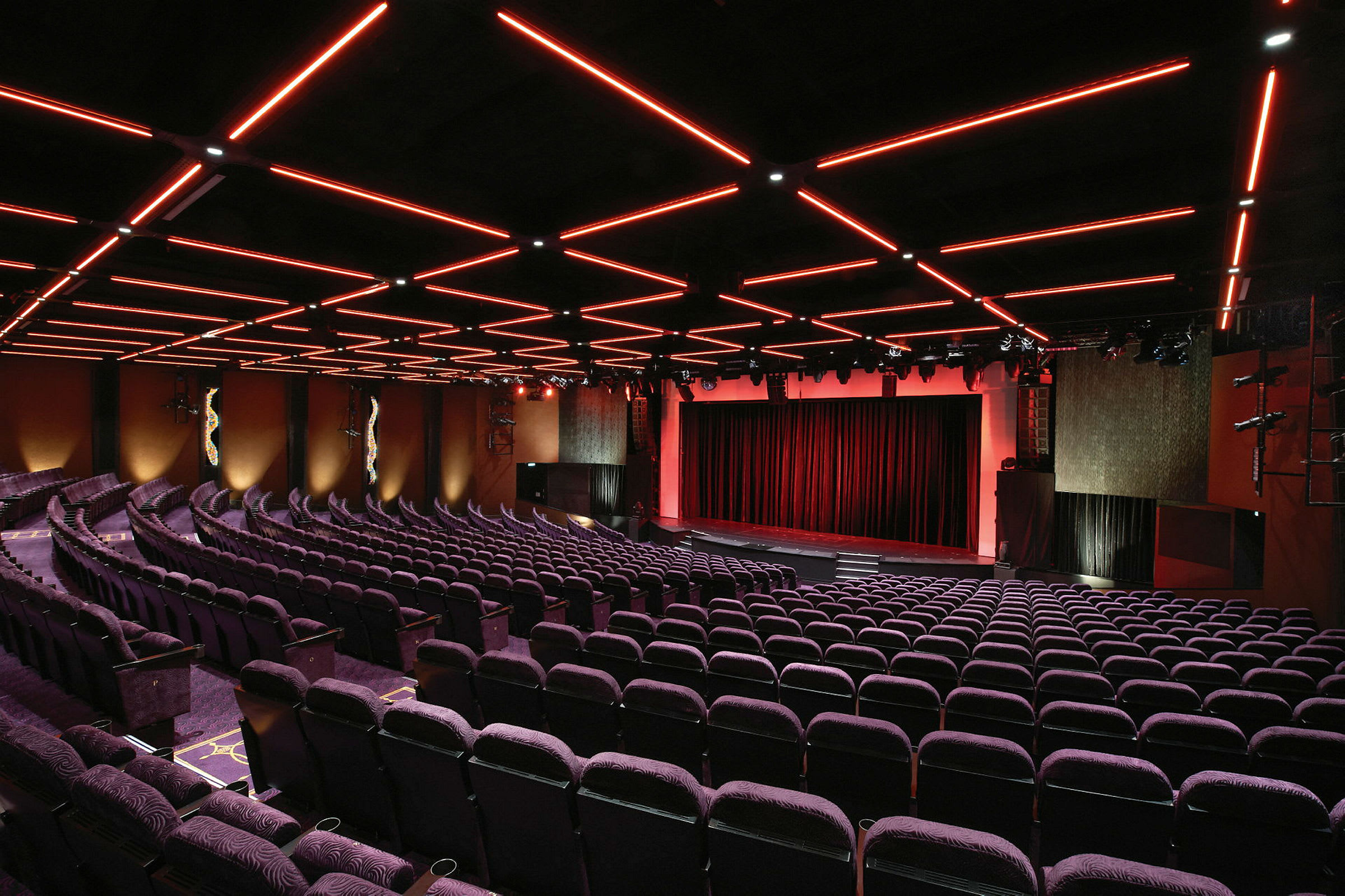 En teatersal med rader av lila stolar och ett upplyst tak med röda streck. Scenen är klädd med röda draperier. (AI-genererad text)