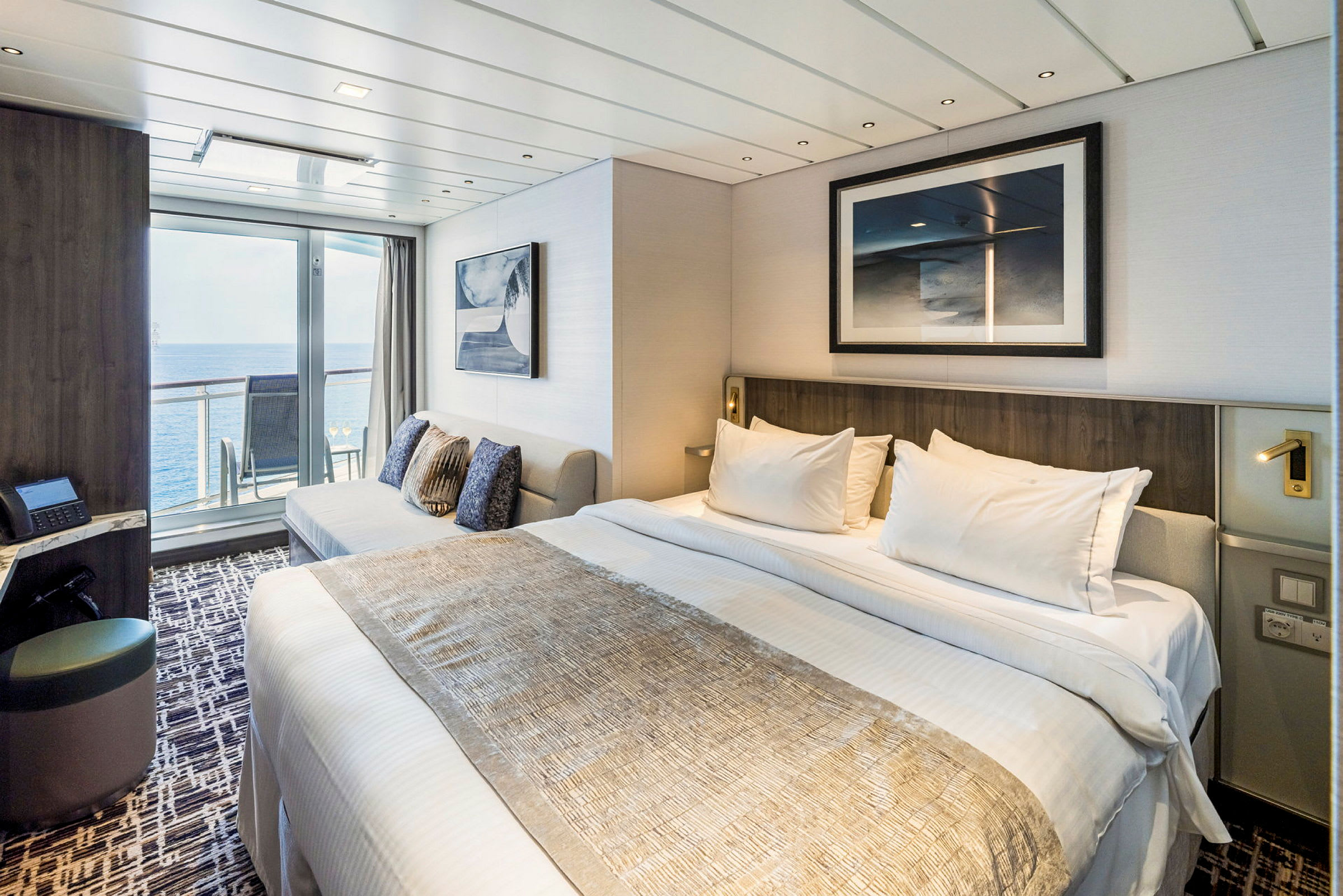 Ett modernt hotellrum med en bäddad säng, en soffa och glasdörrar som leder till en balkong med utsikt över havet. (AI-genererad text)