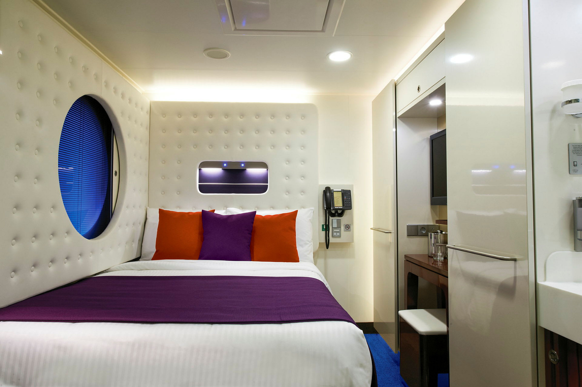 Ett modernt hotellrum med en säng som har lila och orange kuddar. (AI-genererad text)