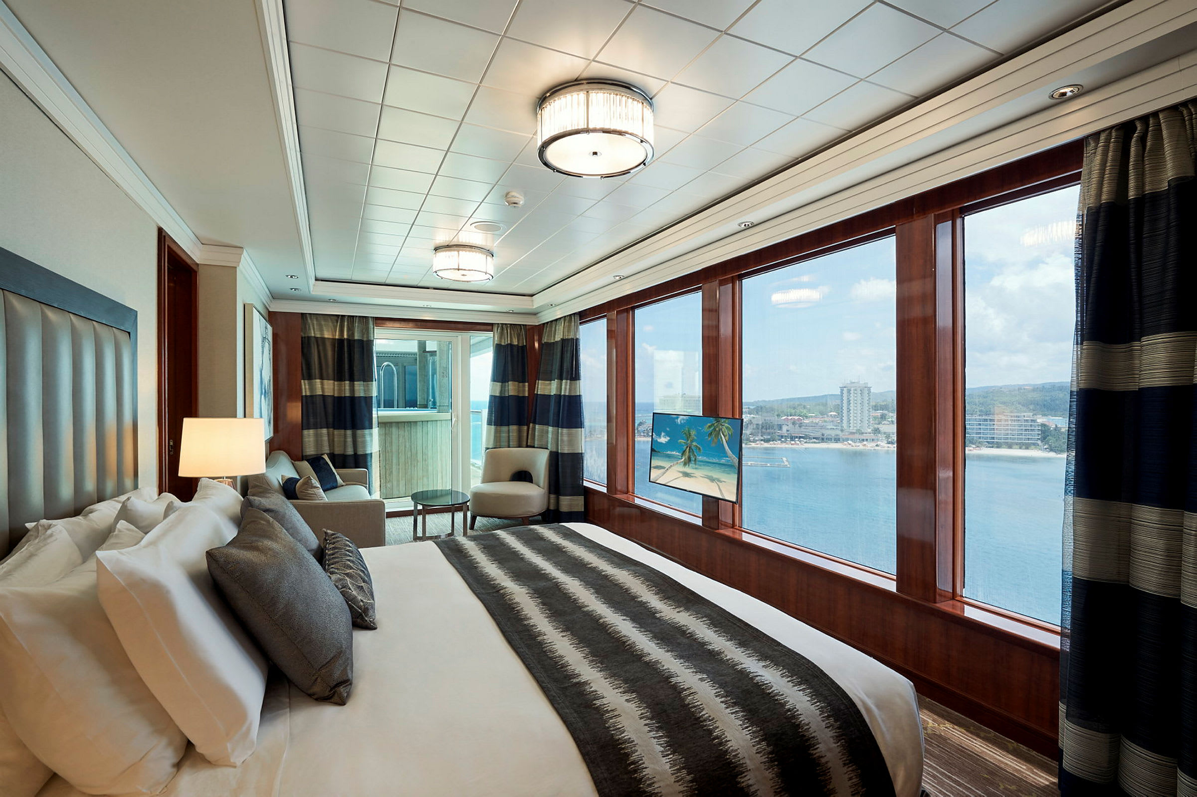 Ett elegant hotellrum med en stor säng, två fåtöljer och stora fönster som visar utsikt över vattnet och en kustlinje. (AI-genererad text)