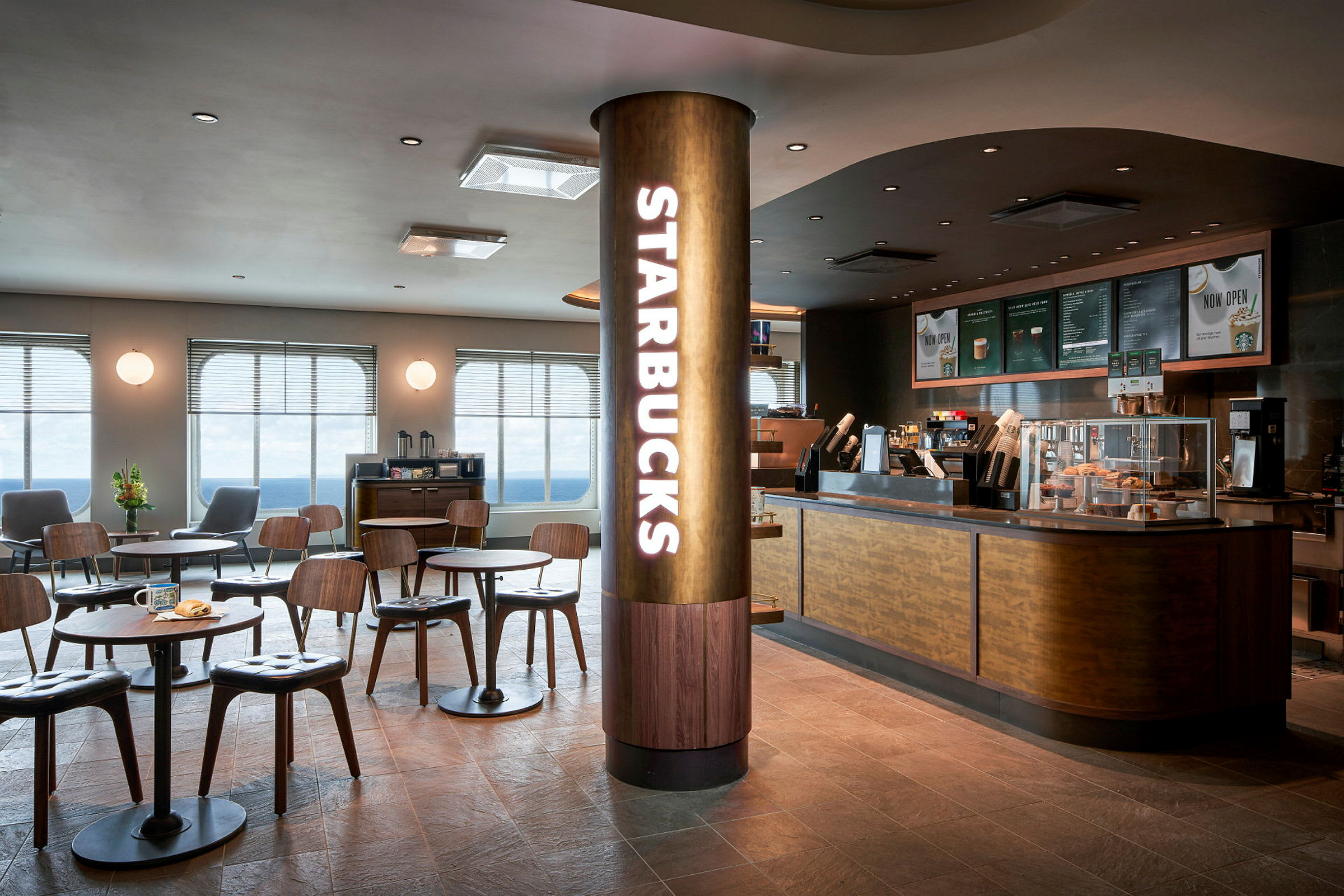 Ett modernt kafé med Starbucks-logotyp på en pelare och sittplatser med bord framför en serveringsdisk. (AI-genererad text)