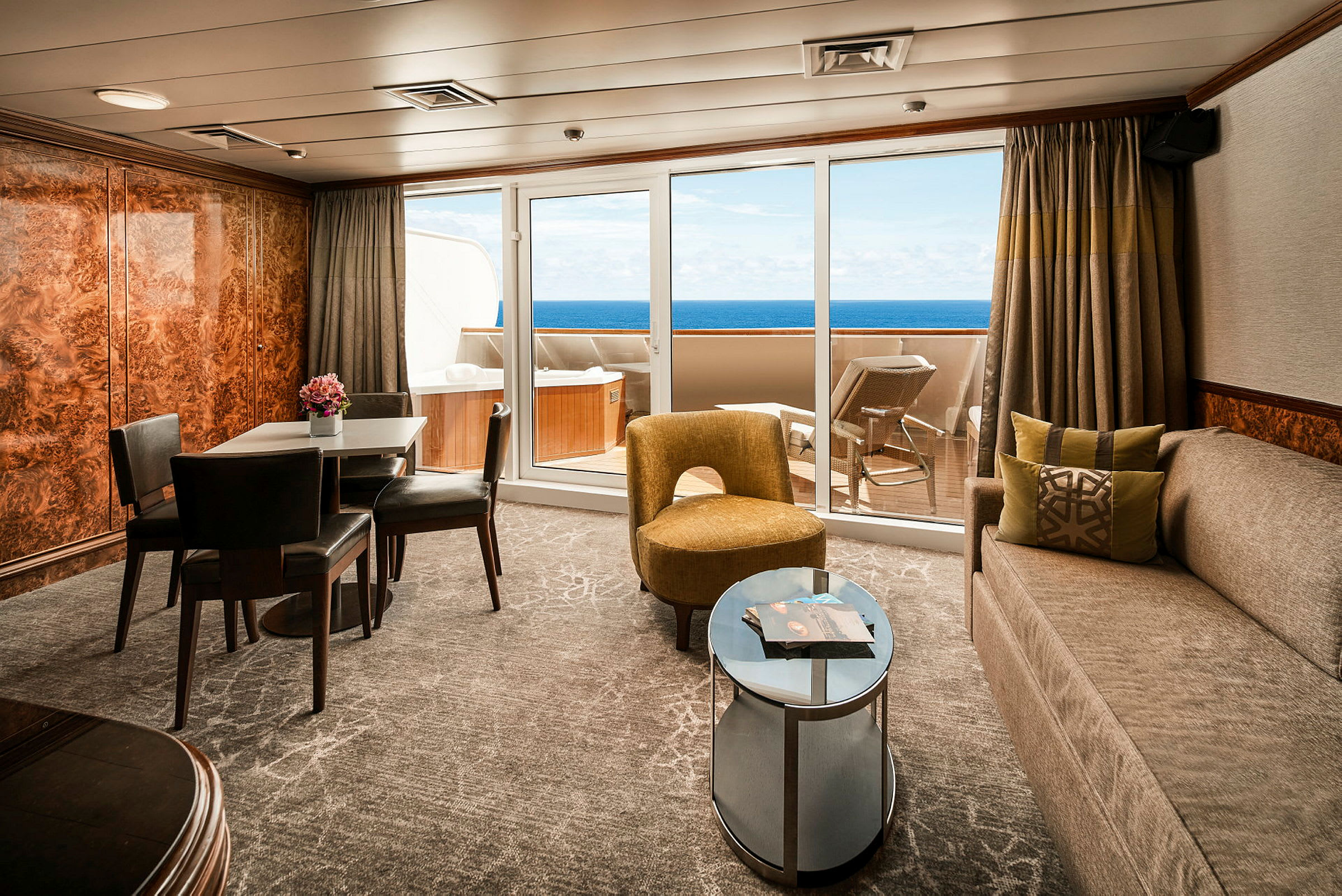Ett möblerat vardagsrum med matbord och stolar, soffa, fåtölj samt utsikt mot en balkong med solsängar och havet i bakgrunden. (AI-genererad text)
