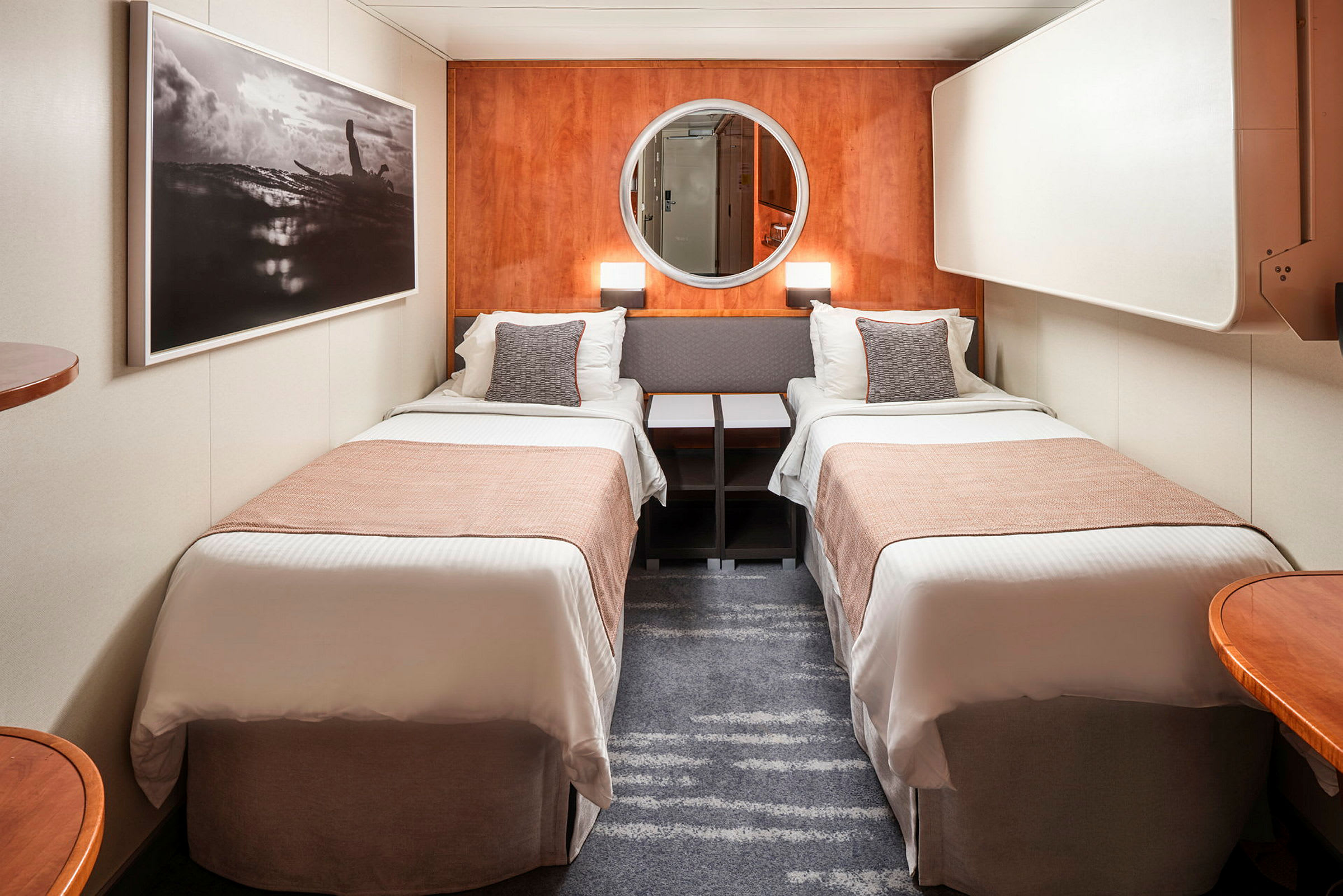 Ett hotellrum med två enkelsängar separerade av ett nattduksbord. Väggarna är dekorerade med en stor tavla och en rund spegel. (AI-genererad text)