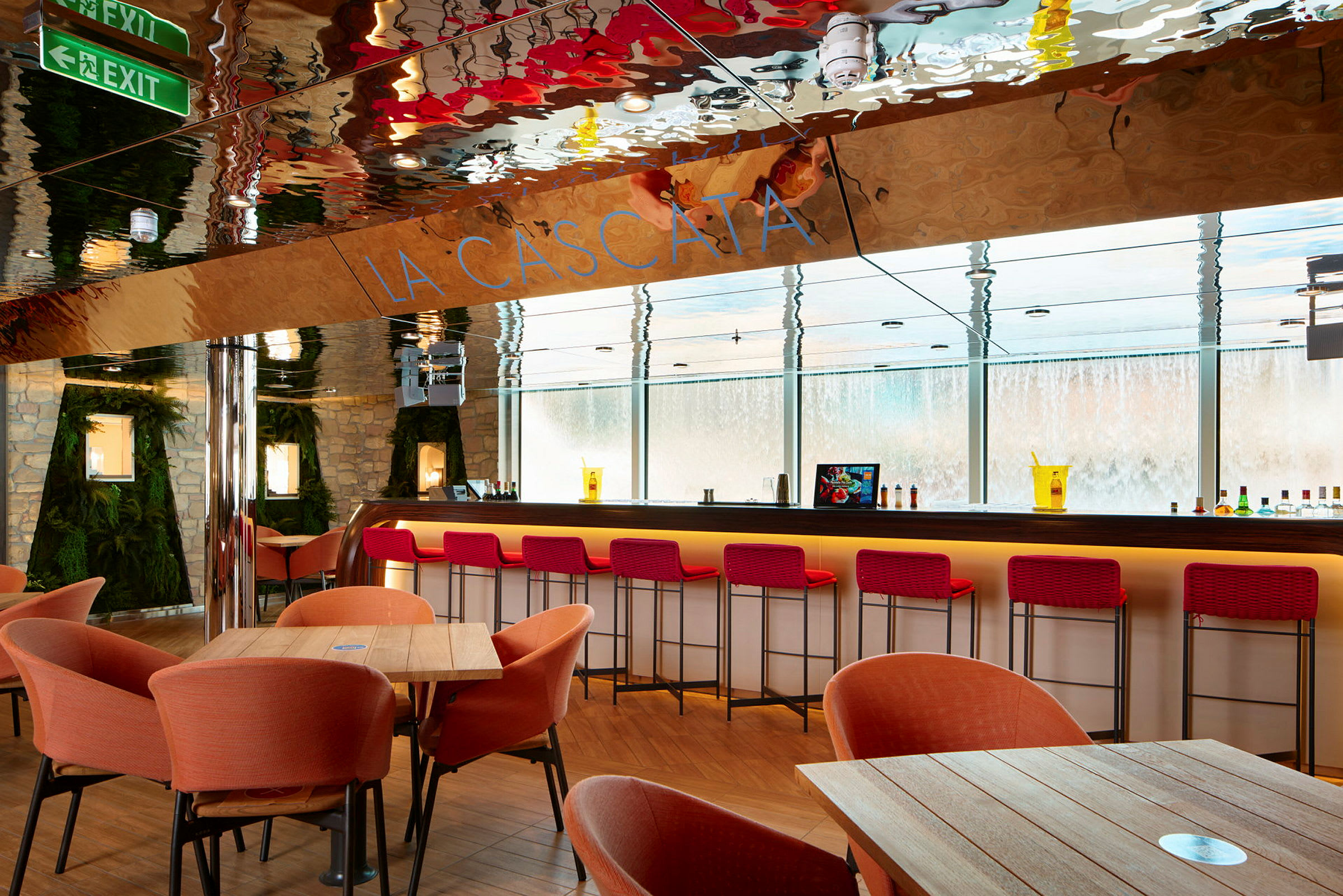 En modern bar med röda barstolar, orangea fåtöljer och träbord. Väggarna är dekorerade med sten och grönska. Glasfönster visar en vatteninstallation utanför. (AI-genererad text)