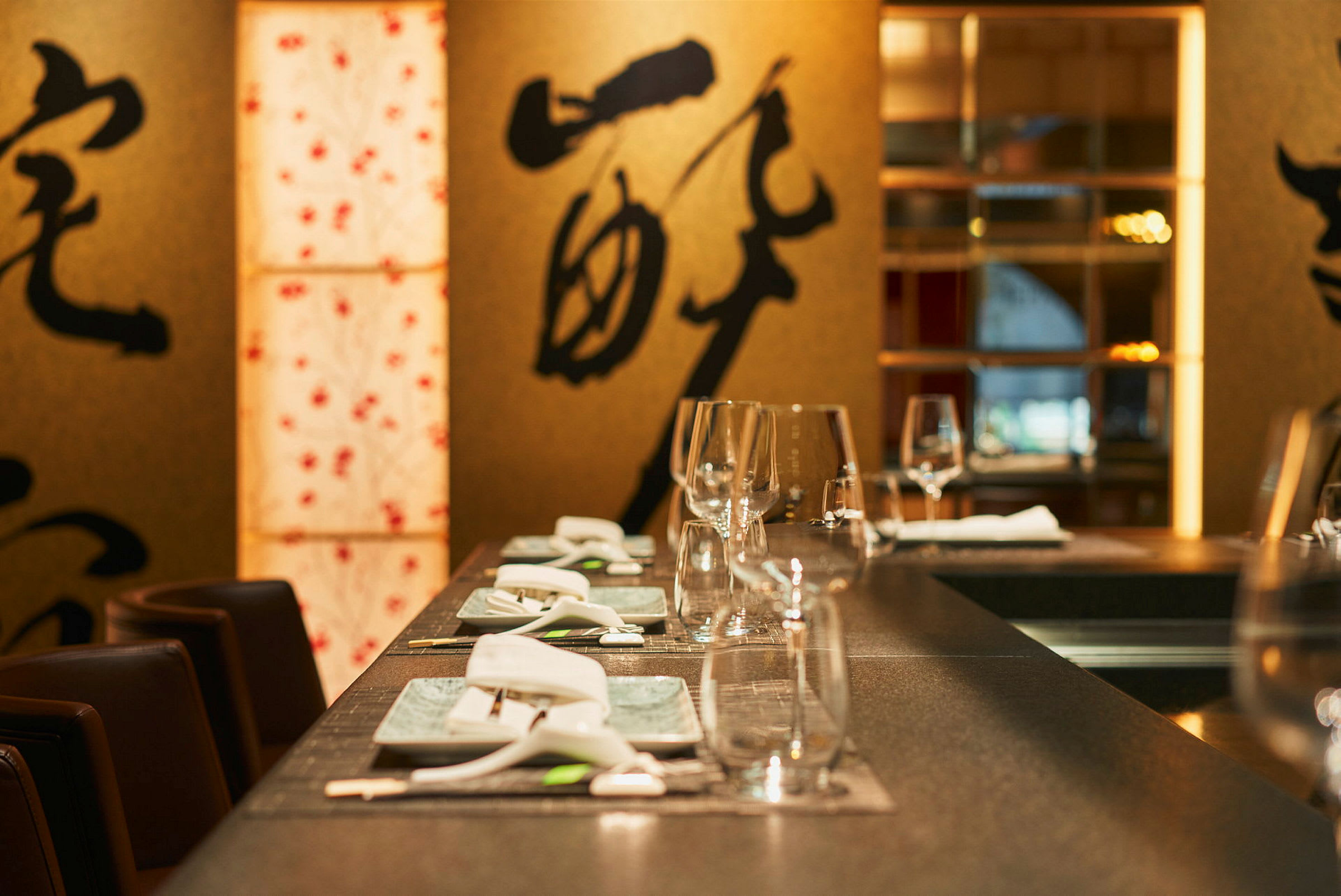 Ett dukat bord på en restaurang, dekorerat med glas, porslin och bestick. I bakgrunden syns dekorationer med asiatiska motiv. (AI-genererad text)