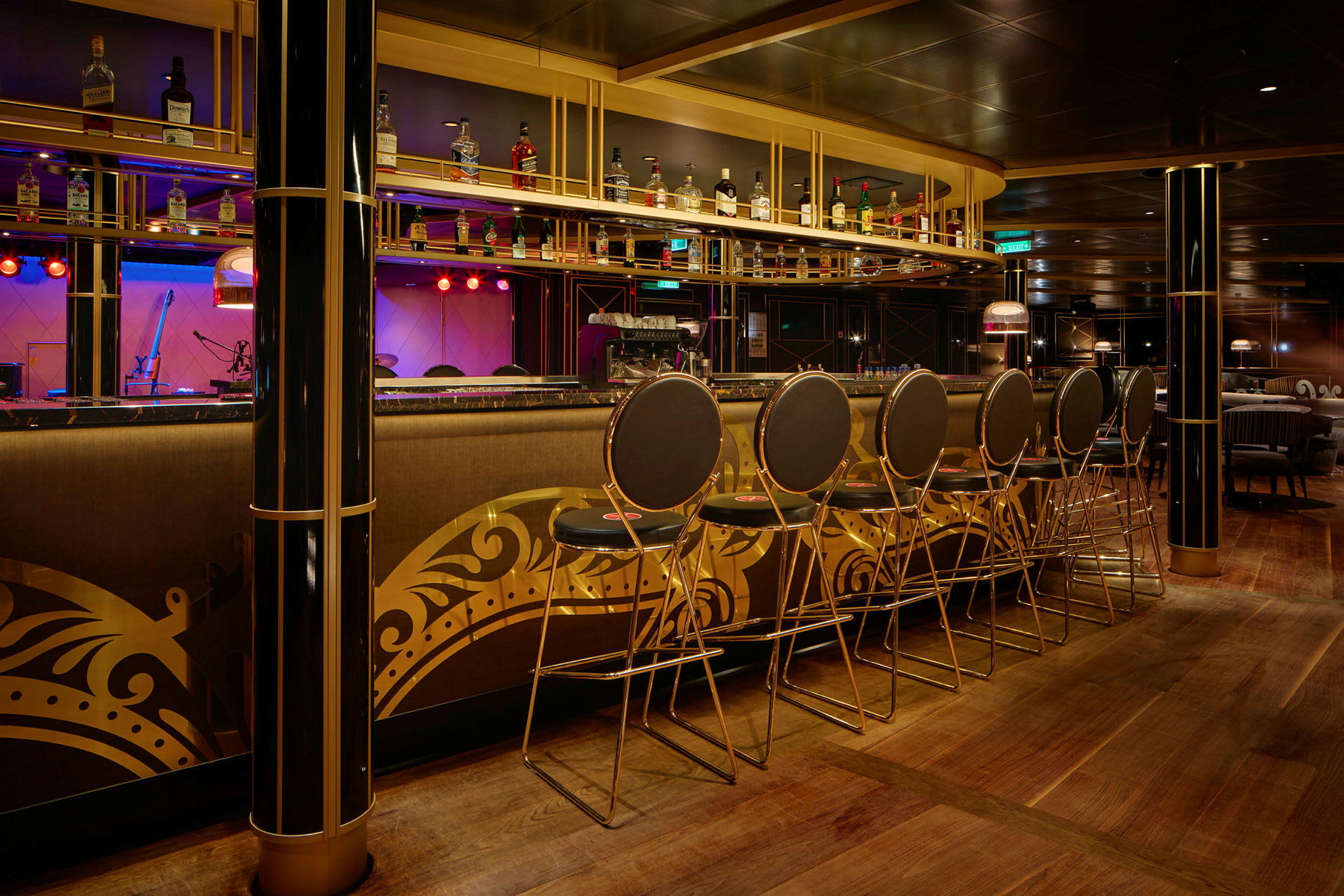 En elegant bar med höga stolar och en hylla med flaskor ovanför bardisken. Interiören har gyllene dekorationer. (AI-genererad text)