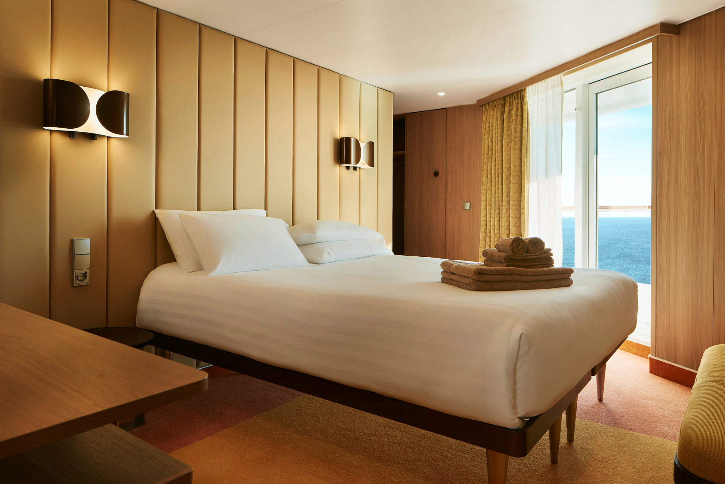 Ett hotellrum med en bäddad säng, handdukar på sängen, och havsutsikt genom glasdörren. (AI-genererad text)