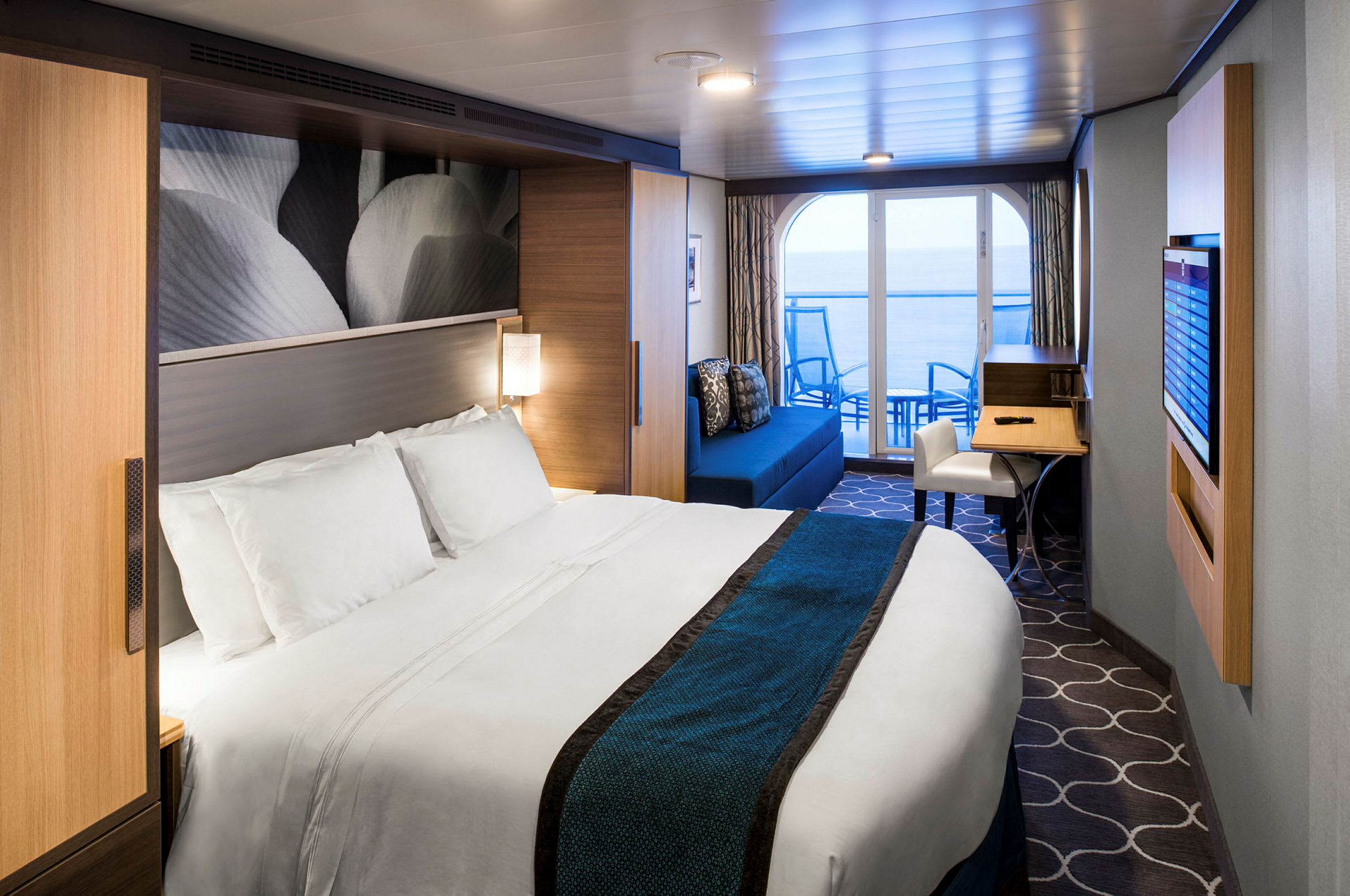 Ett modernt hotellrum med en stor säng, utsmyckad med vita och blå sängkläder. Rummet har en sittgrupp och skrivbord, samt en balkong som vetter mot havet. (AI-genererad text)