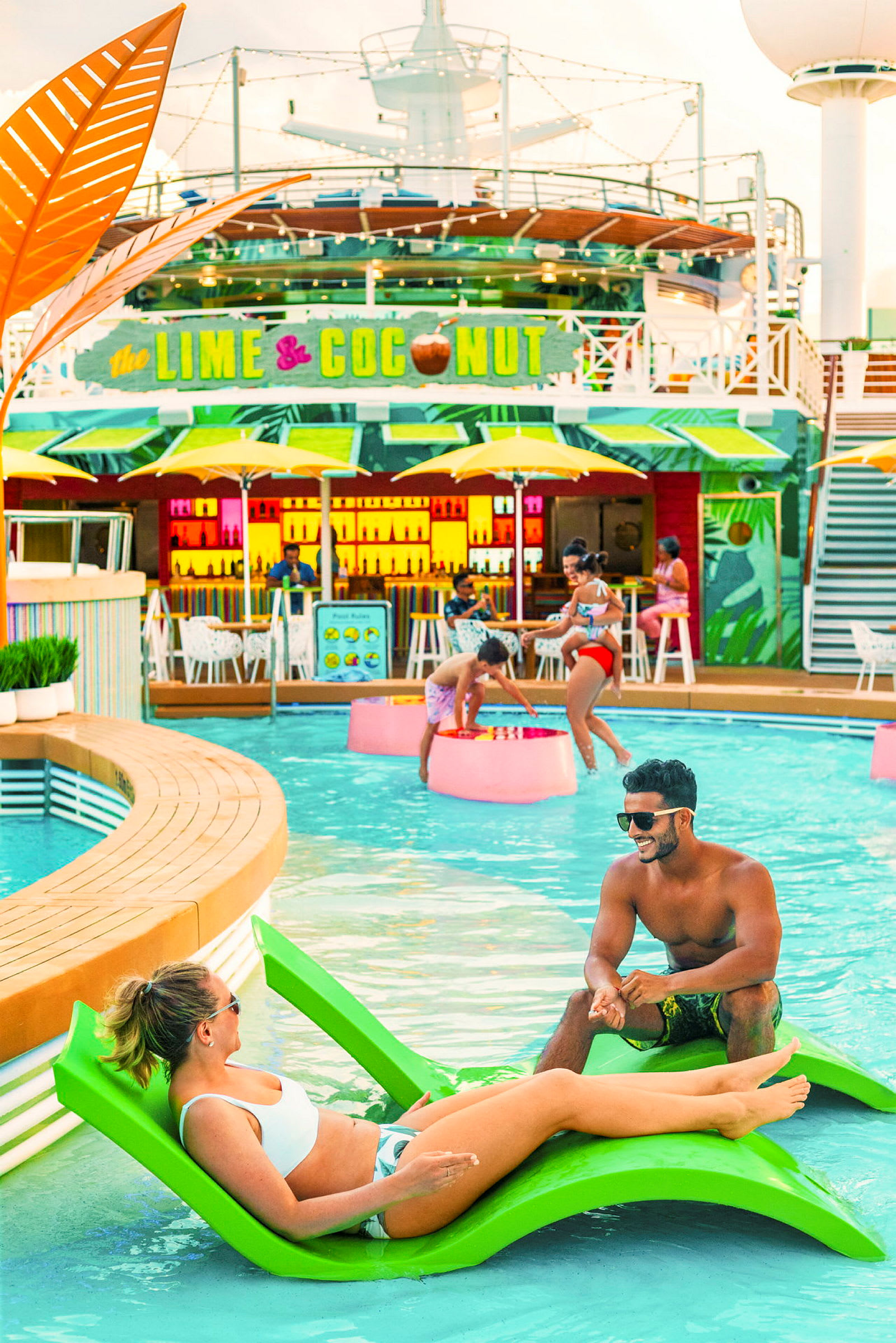 En pool omgiven av människor som njuter av solen och simmar, med en färgglad bar i bakgrunden. (AI-genererad text)