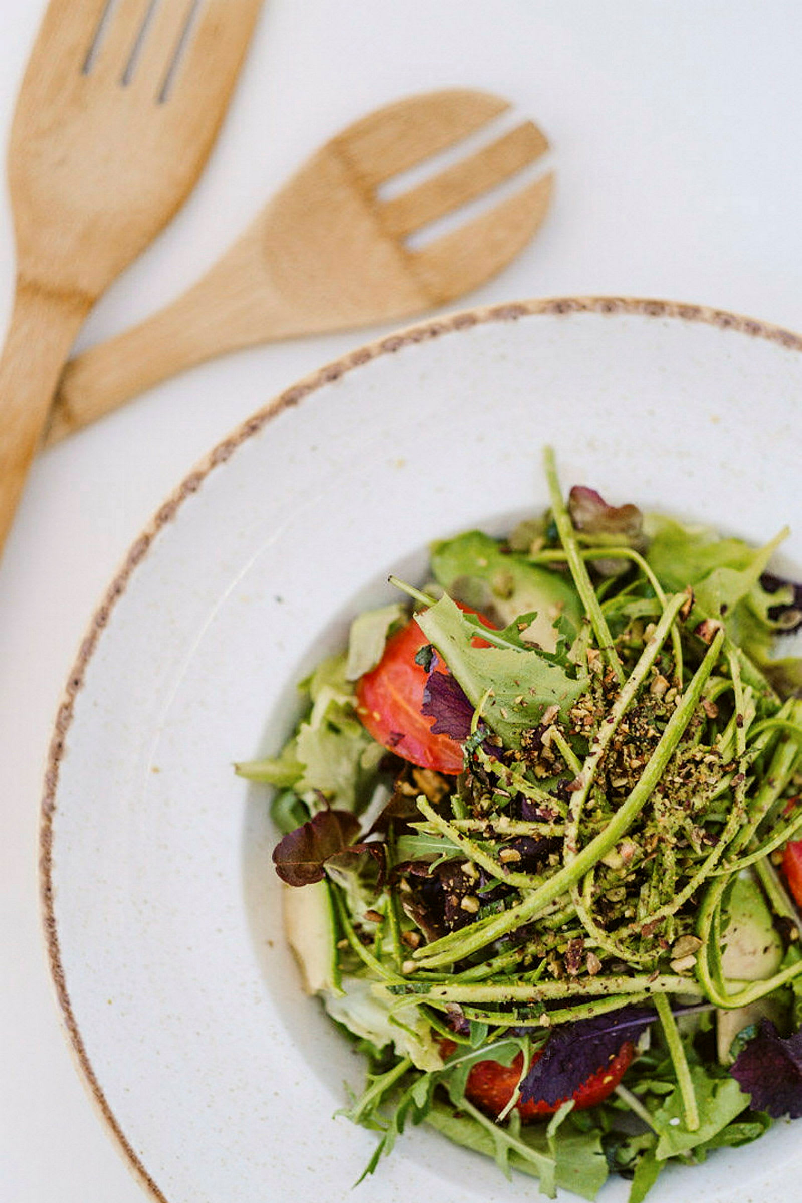En tallrik med en färgglad sallad som innehåller bladgrönt, tomater och avokado, med grovhackade nötter på toppen. Två träbestick ligger bredvid. (AI-genererad text)