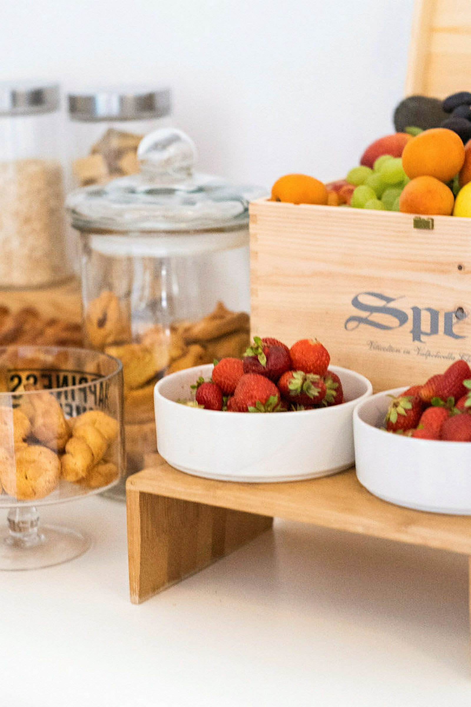 Bild av en matuppställning med färska jordgubbar i vita skålar, en trälåda med andra frukter samt glasburkar med olika livsmedel. (AI-genererad text)