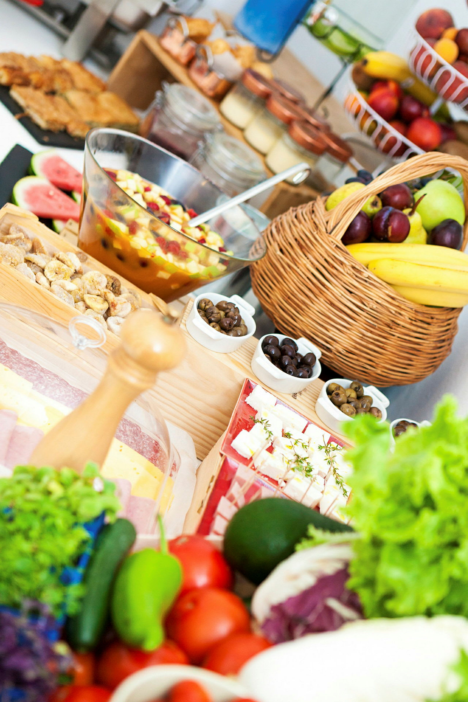 Ett bord fyllt med olika färska frukter, grönsaker och mat, inklusive en fruktsallad i en glasbehållare och ett brödkorg med bananer och plommon. (AI-genererad text)