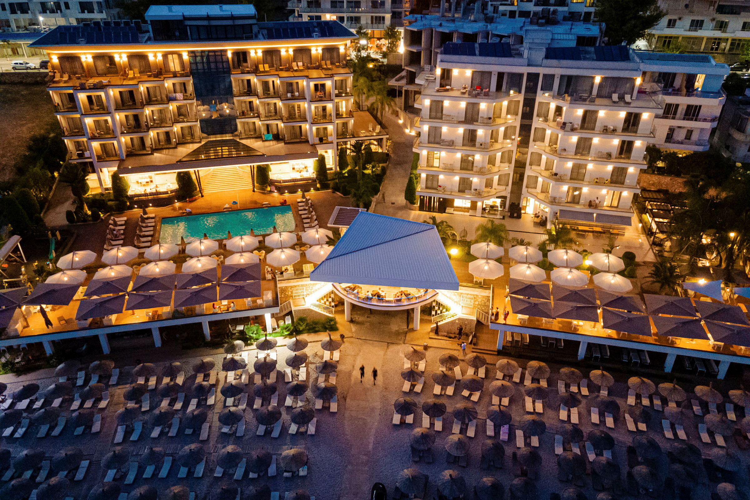 Flygvyn av en upplyst hotellanläggning på kvällen, med pool och solstolar nära stranden. (AI-genererad text)