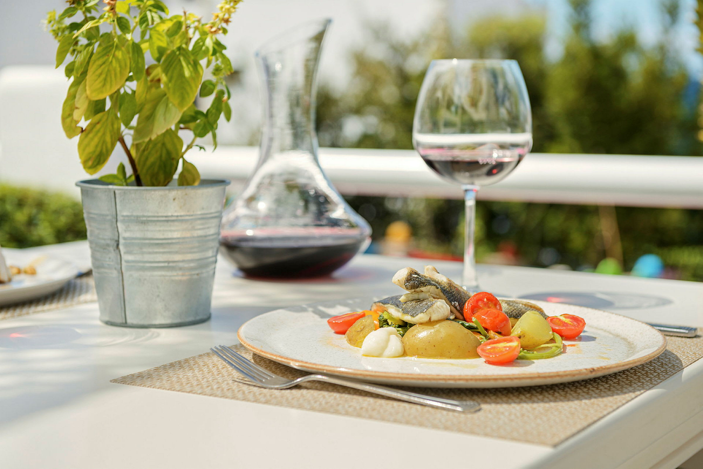 En tallrik med mat, bestående av fisk, potatis och körsbärstomater, står på ett bord tillsammans med ett vinglas, en karaff med vin och en krukväxt. (AI-genererad text)