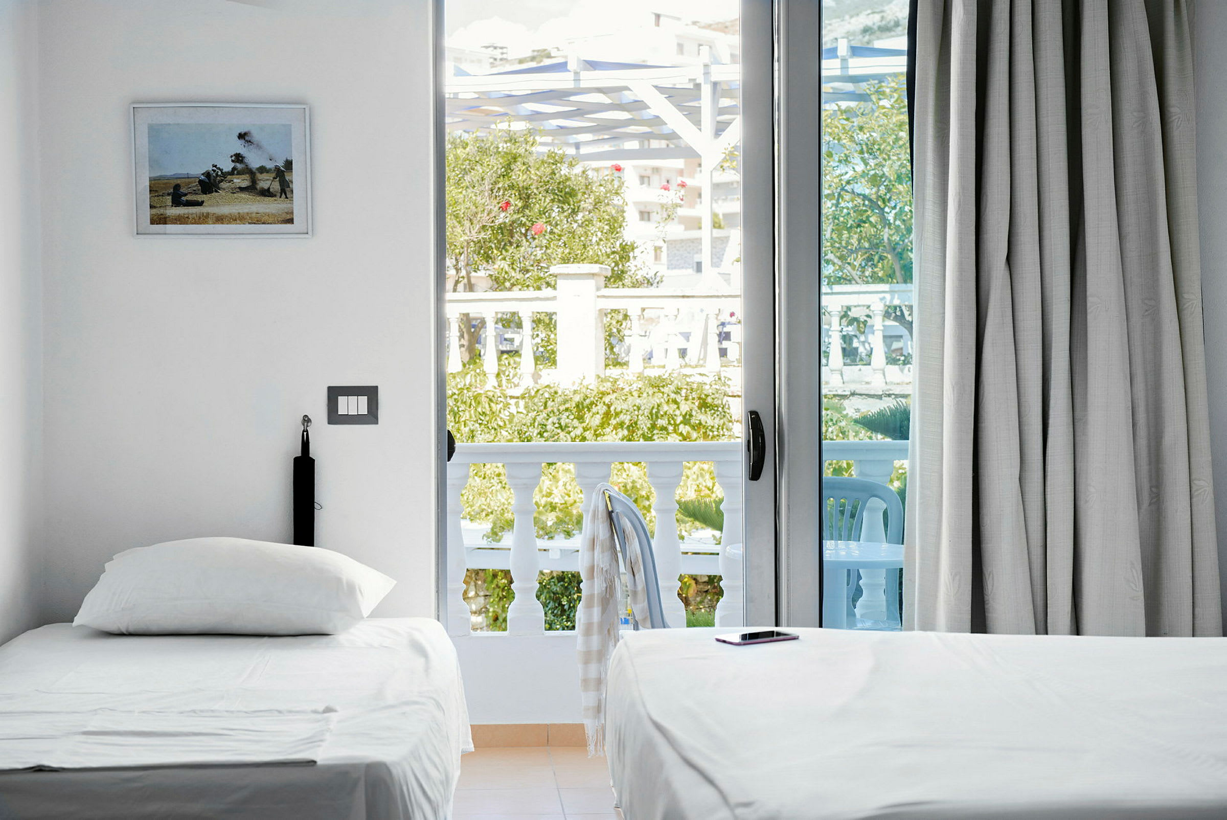 En ljus hotellrum med två enkelsängar, en balkong med utsikt mot grönska och utemöbler. (AI-genererad text)
