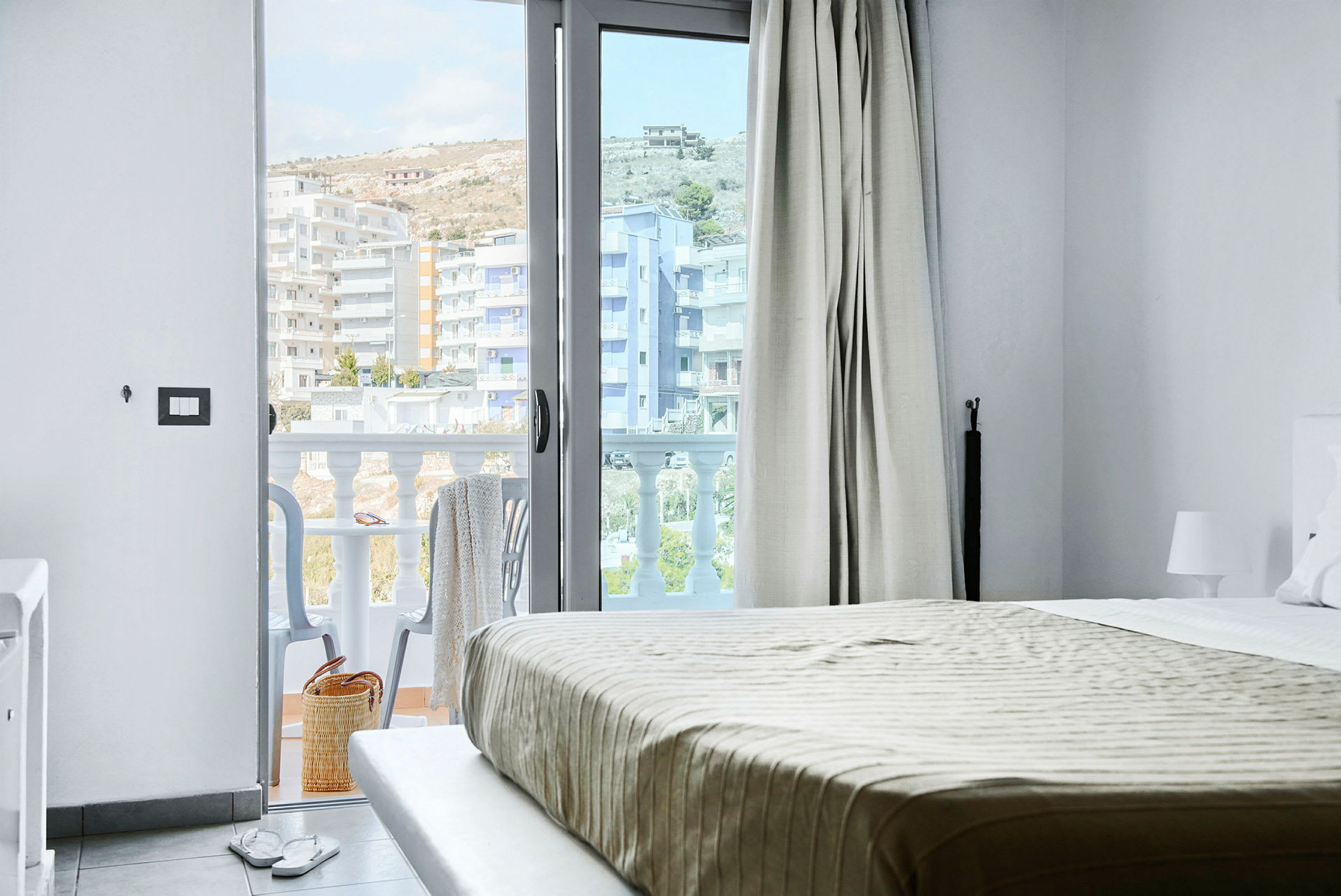 Utsikt från ett hotellrum, med säng och en öppen balkongdörr. Balkong med stolar och utsikt över byggnader. (AI-genererad text)