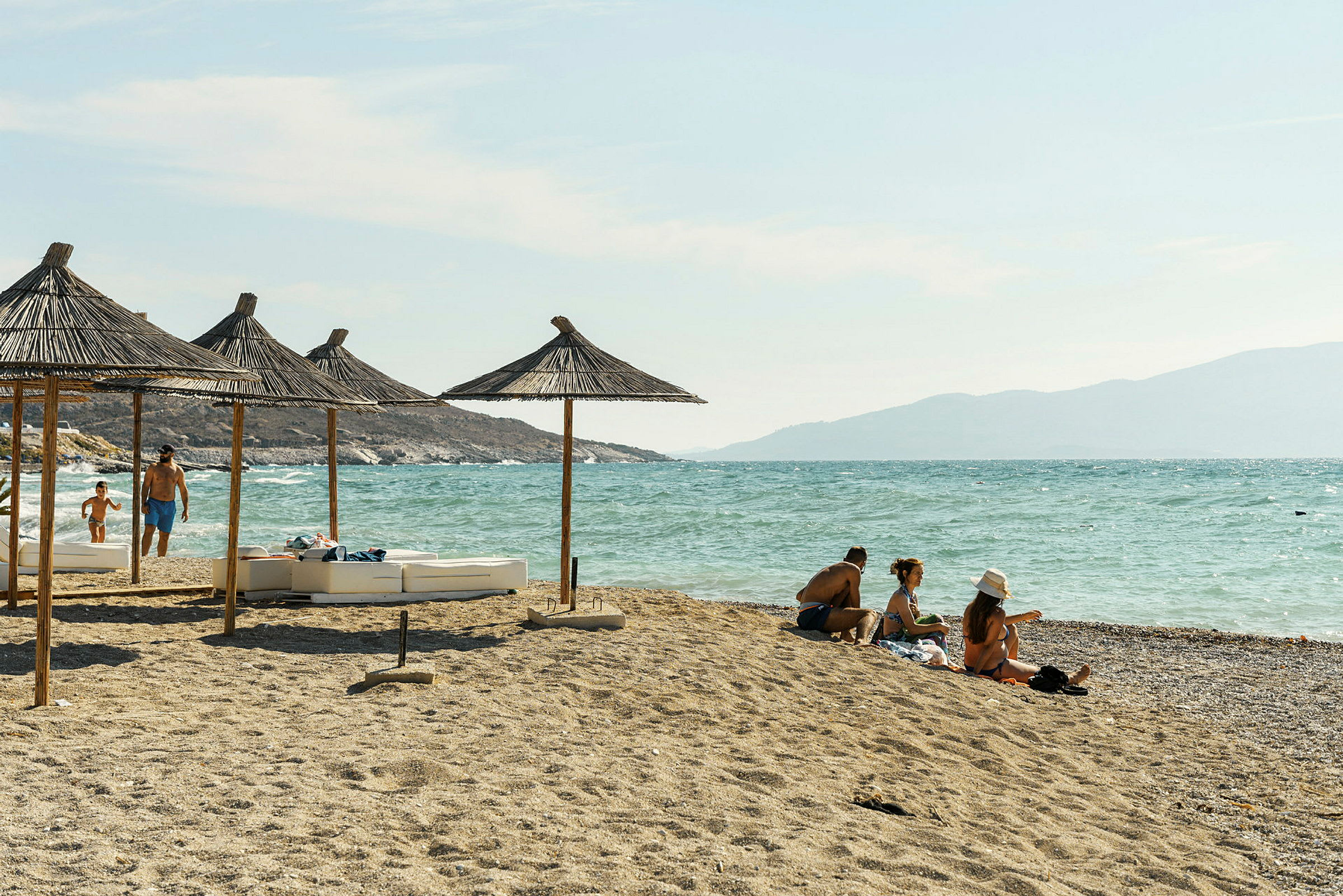 Strand med parasoller av halmtak och människor som njuter av solen vid havet. (AI-genererad text)