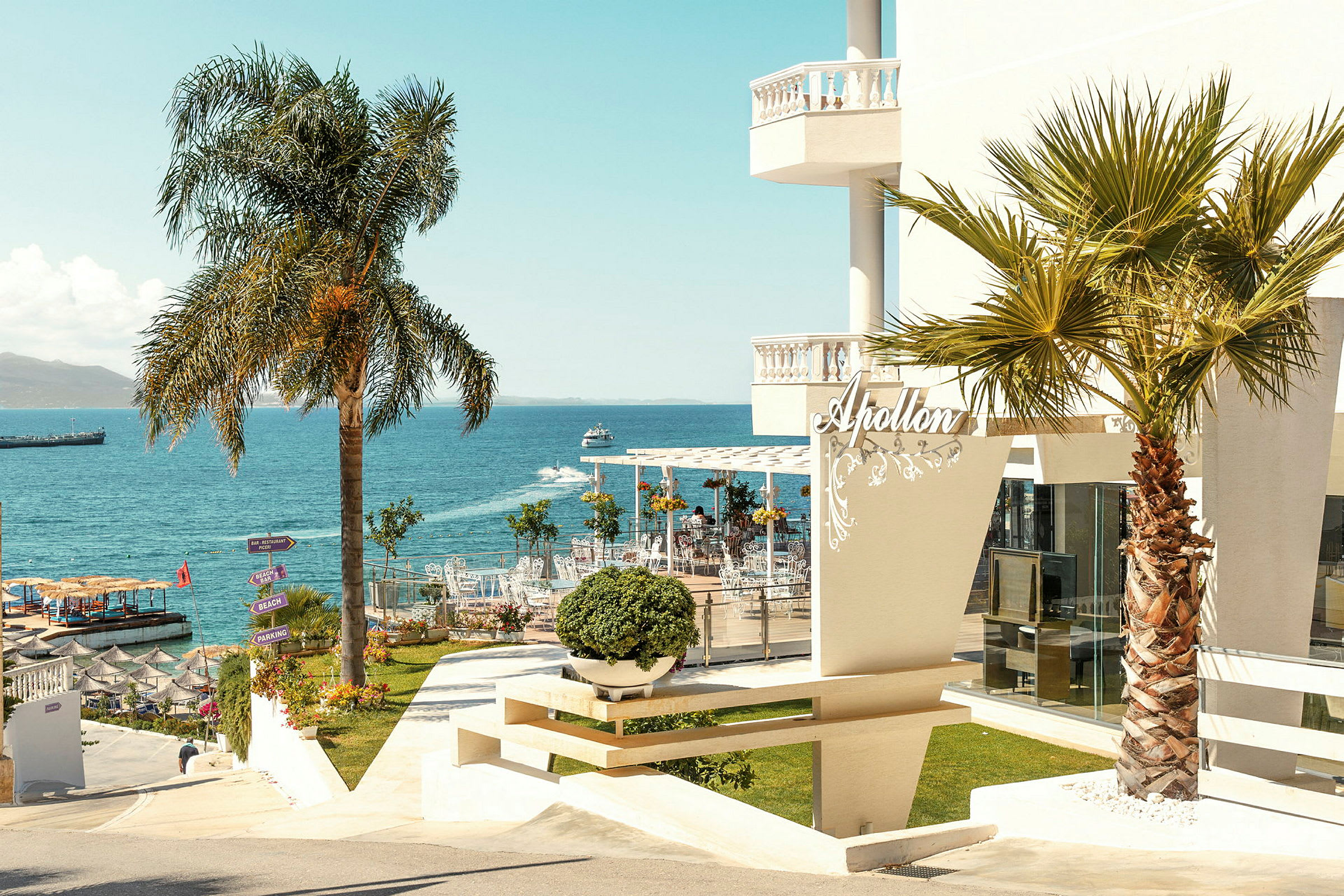 Hotellbyggnad med palmer i förgrunden och havet i bakgrunden. (AI-genererad text)