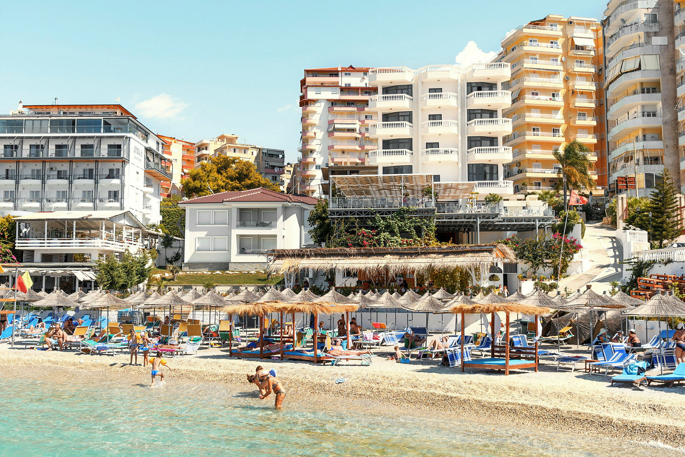 Strand med solstolar och parasoller framför byggnader. (AI-genererad text)