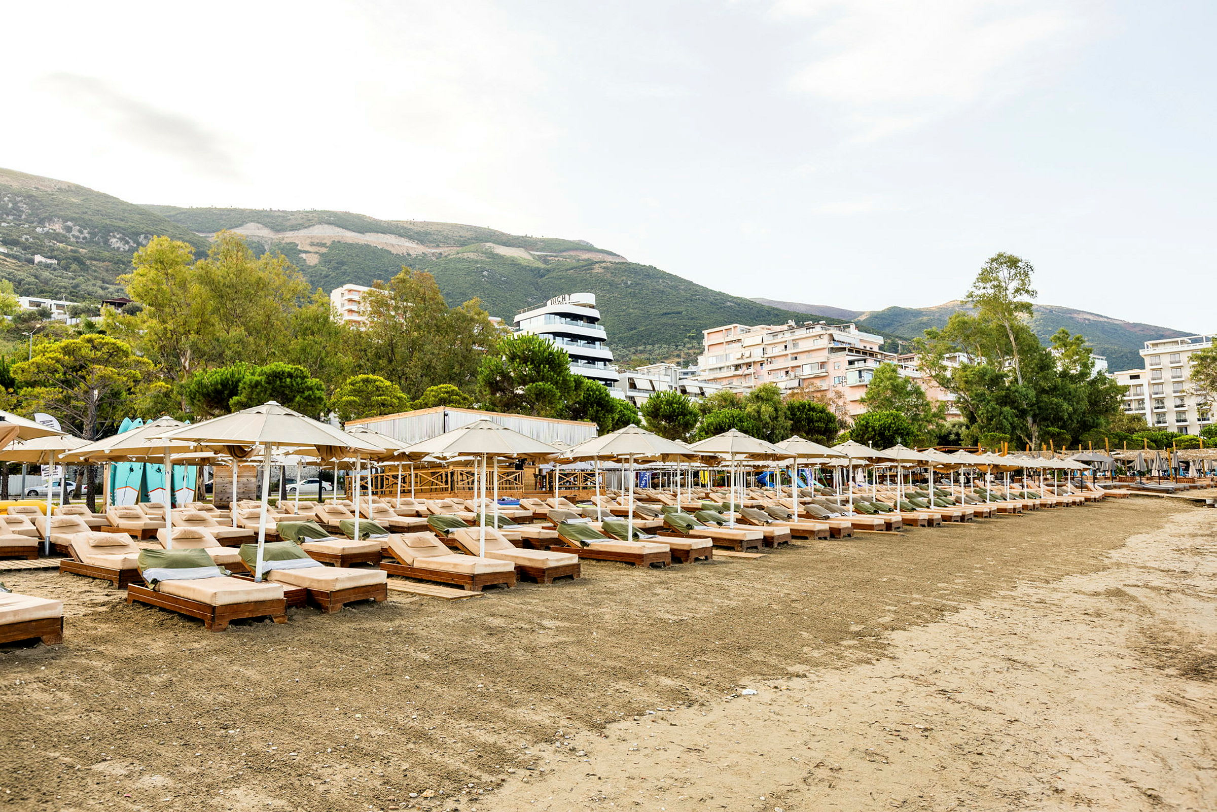 Strand med solstolar och parasoller framför byggnader och gröna berg. (AI-genererad text)