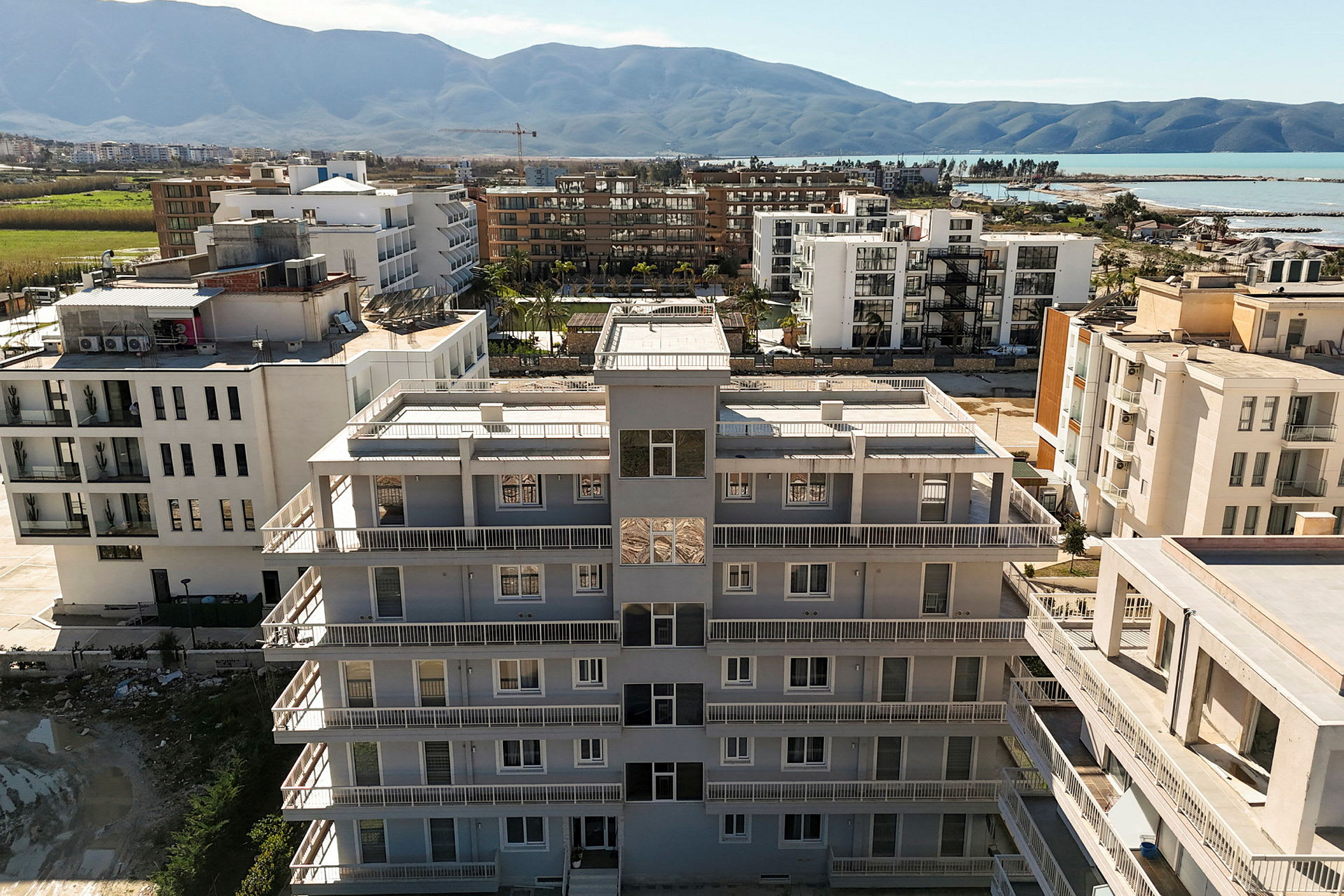 Ett bostadshus med vit fasad och balkonger sett från ovan. I bakgrunden syns fler byggnader, grönområde och berg. (AI-genererad text)
