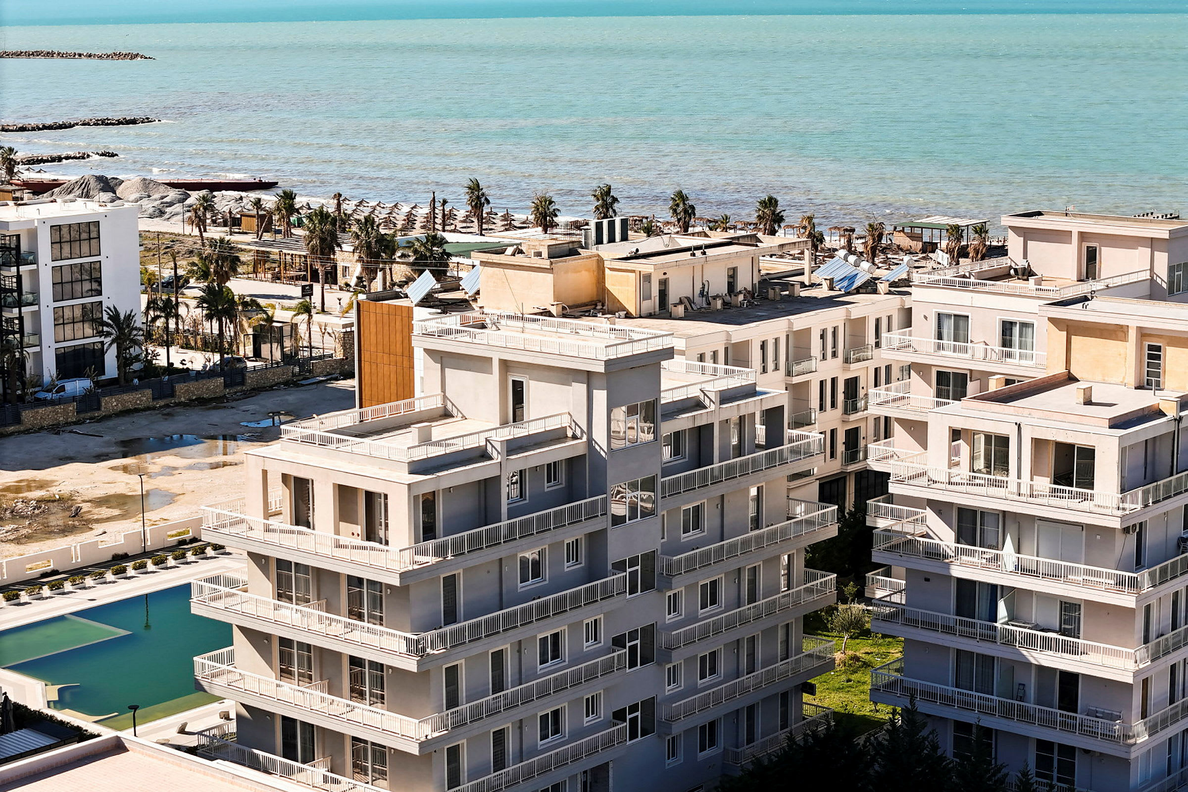 Flera byggnader med balkonger runtom och en pool syns i förgrunden, medan stranden och havet syns i bakgrunden. (AI-genererad text)