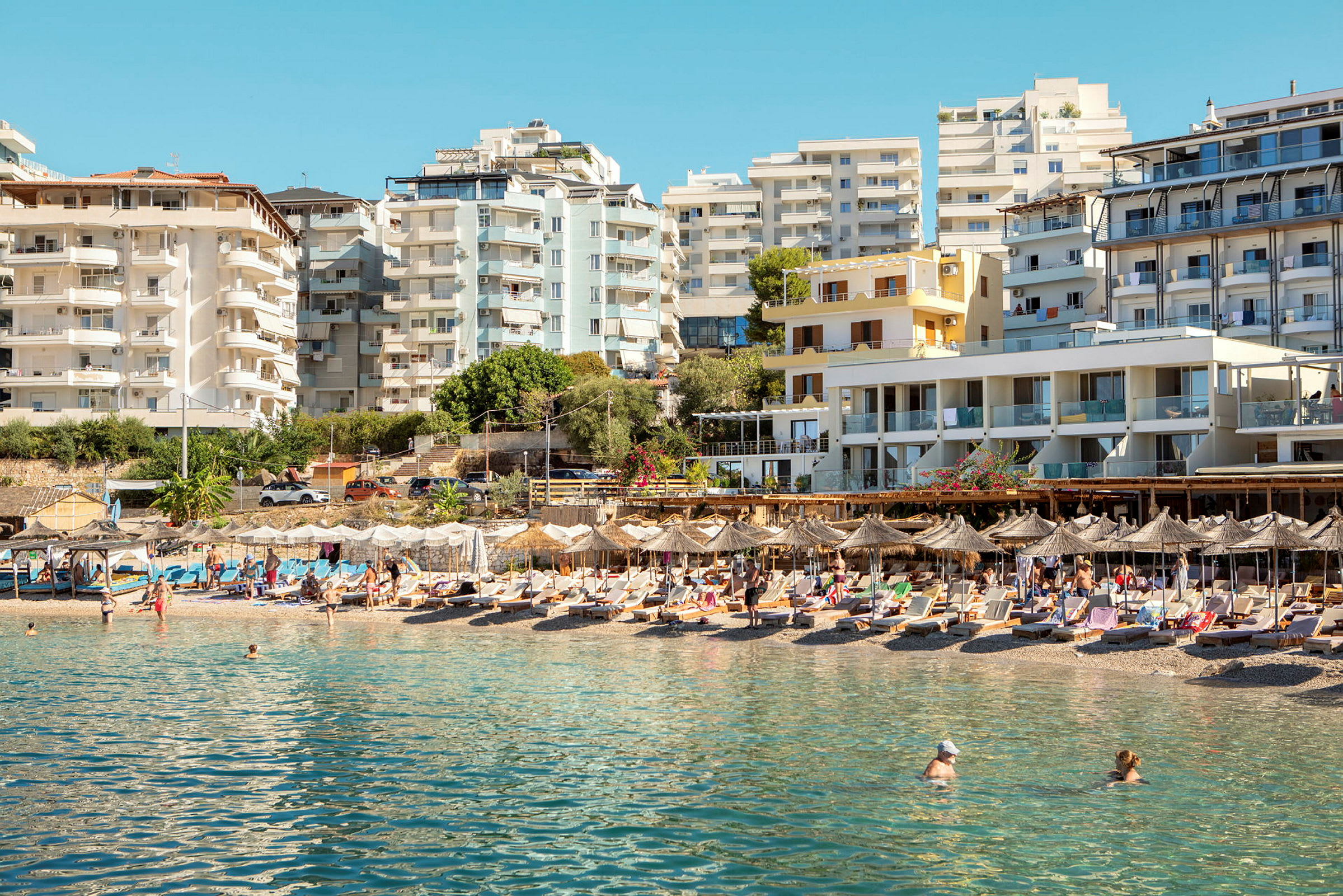 En strand med solstolar och parasoller framför flera modern byggnader. (AI-genererad text)