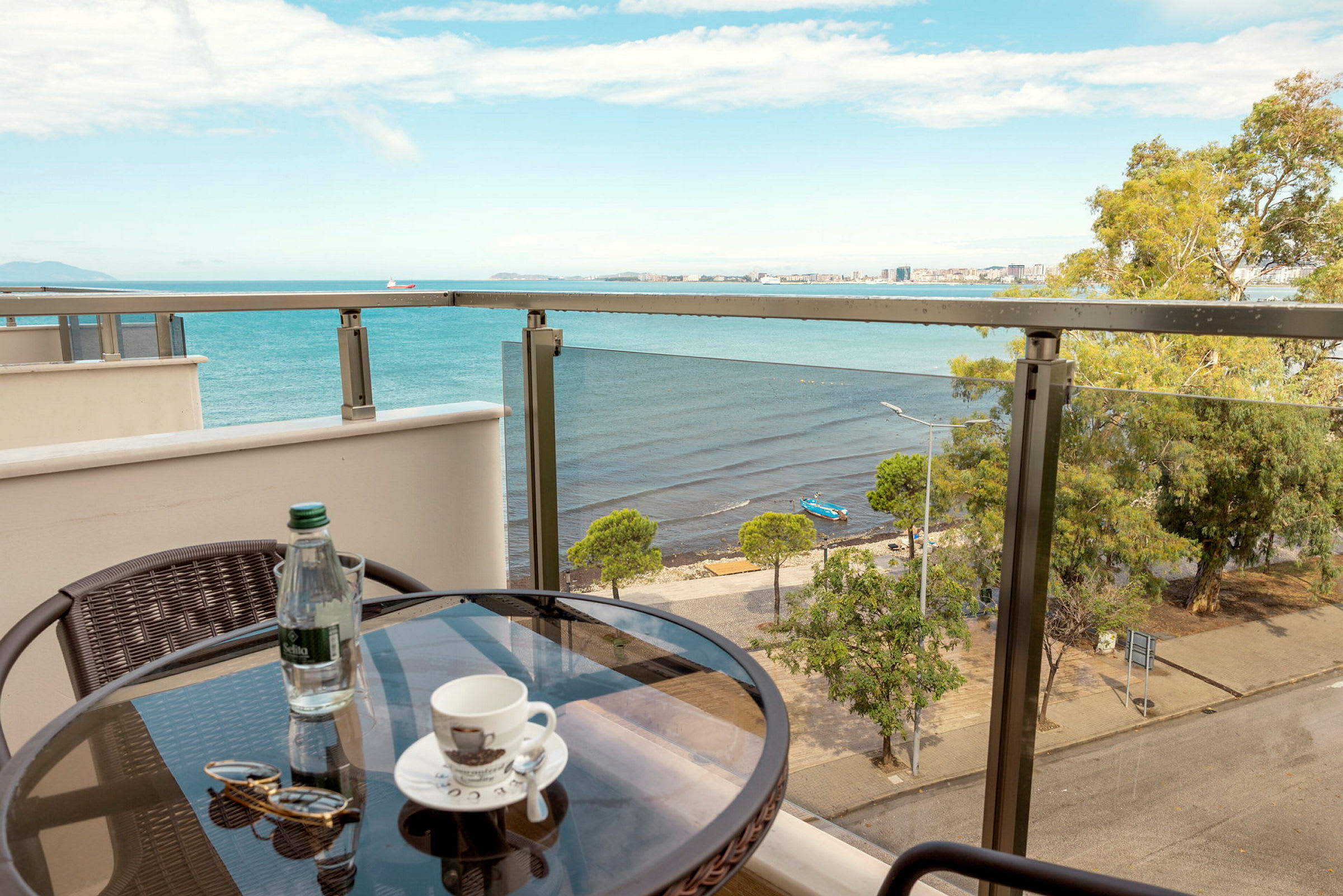 Utsikt från en balkong med ett litet bord där det står en kaffekopp, en flaska vatten och ett par solglasögon. I bakgrunden syns ett hav och en strand med träd. (AI-genererad text)