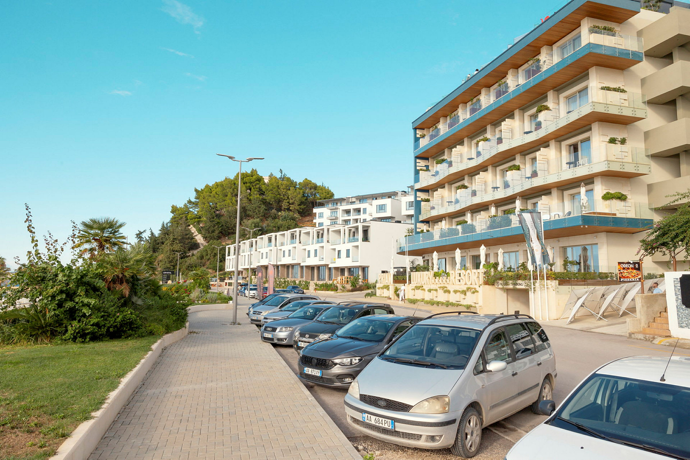 Fotografi av en modern resortbyggnad och parkerade bilar längs en trottoar, med gröna träd och växter i bakgrunden. (AI-genererad text)