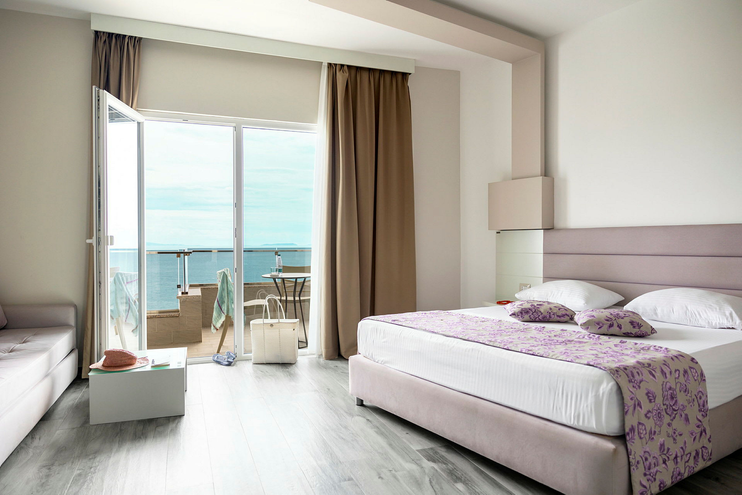 Ett hotellrum med en stor säng, glasdörrar till en balkong och utsikt över havet. (AI-genererad text)