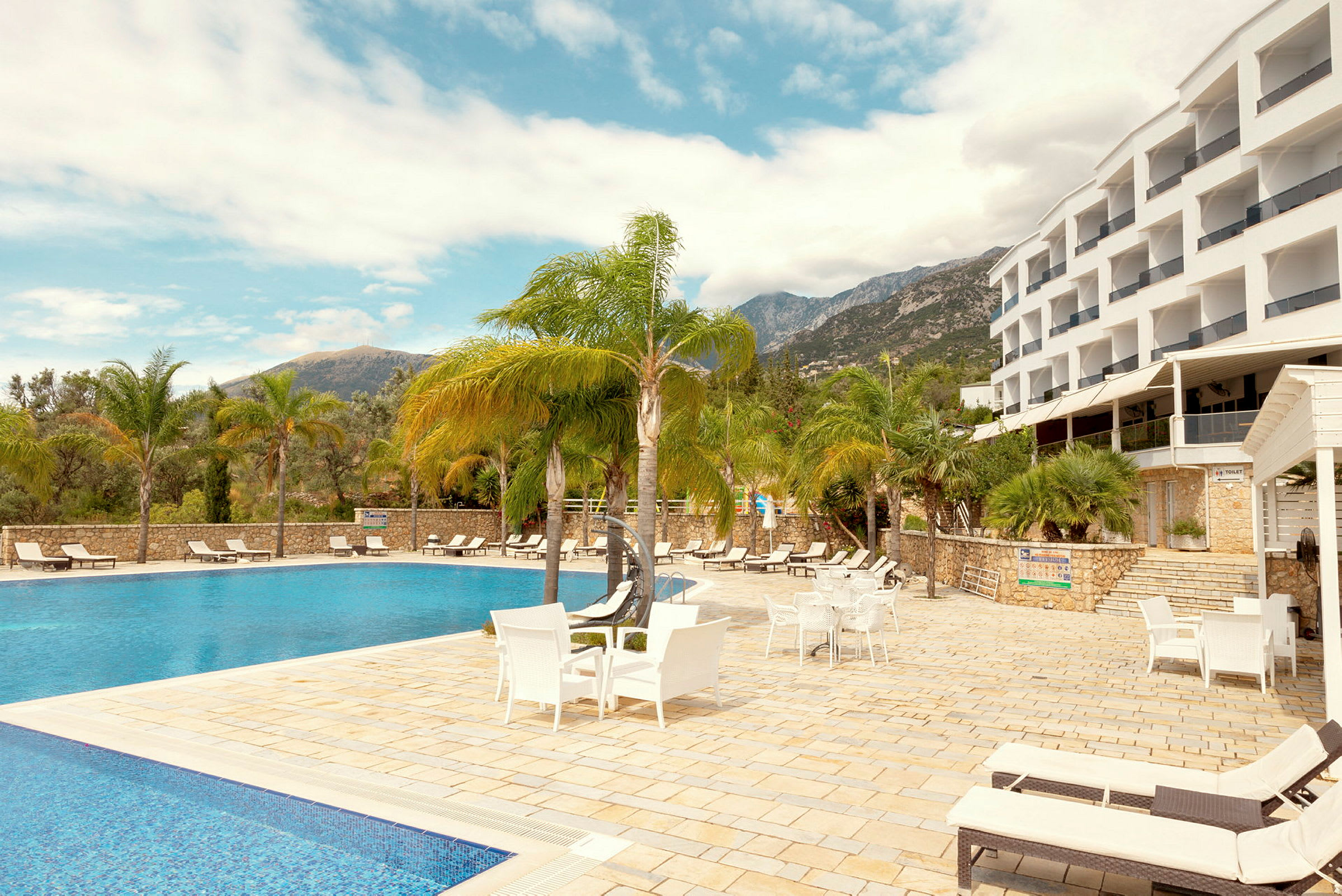 Bild av en hotellpool omgiven av solstolar och palmer, med byggnaden i bakgrunden. (AI-genererad text)