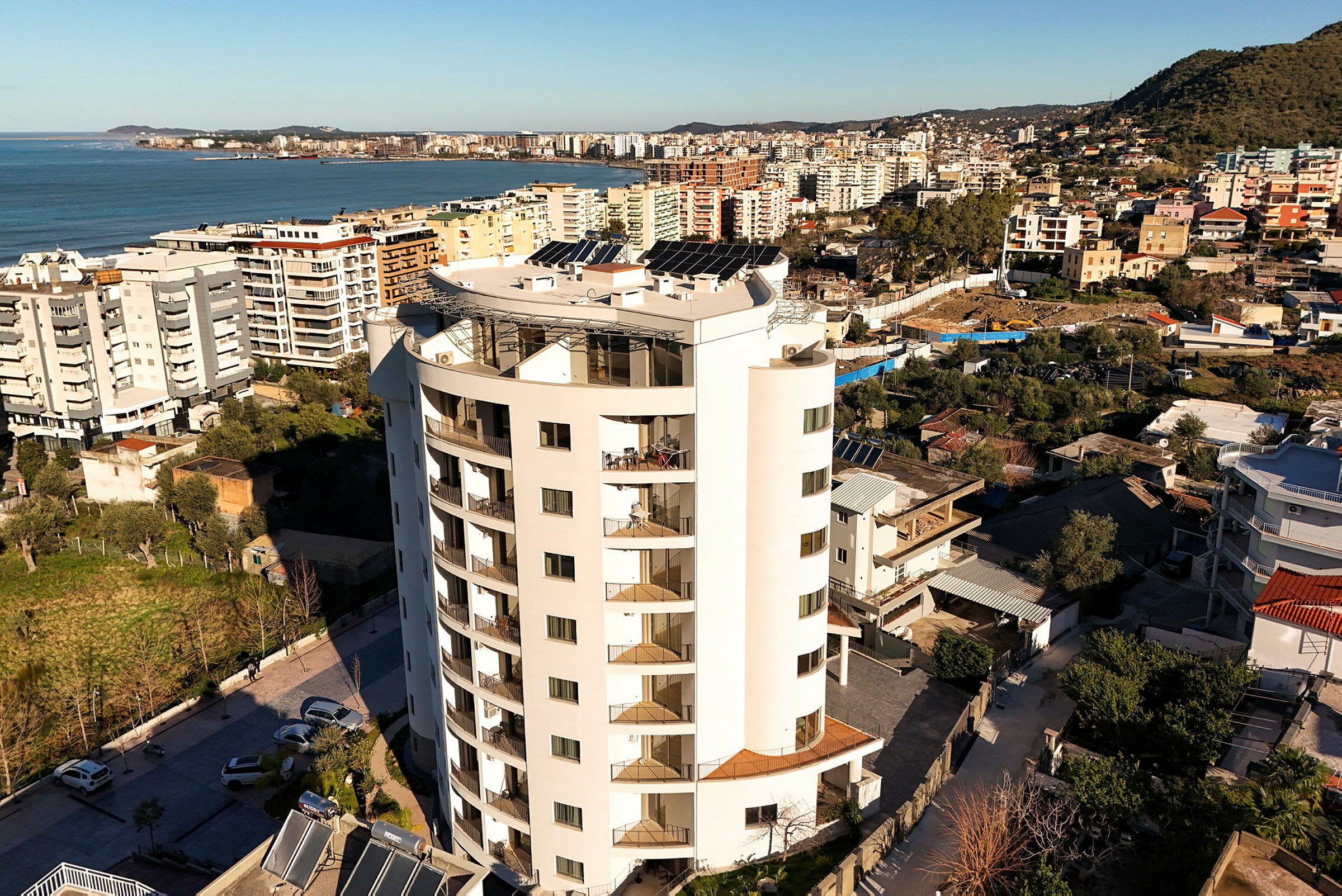 Bild av en vit modern byggnad med flera våningar, omgiven av andra byggnader och med havet i bakgrunden. (AI-genererad text)