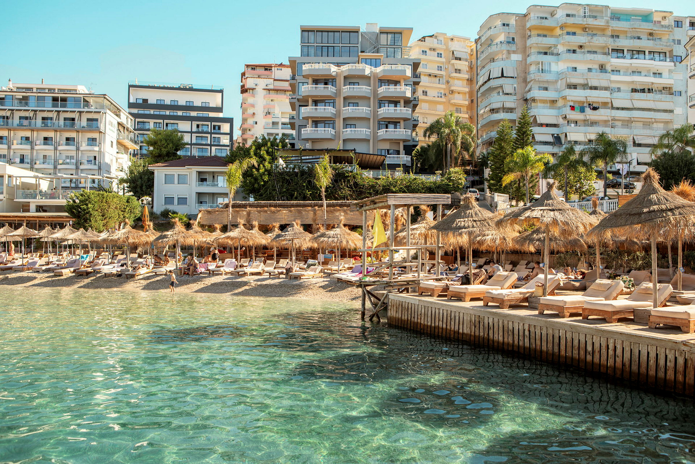 Strand med solstolar och parasoller, omgivna av hotellbyggnader och en klarblå himmel. (AI-genererad text)