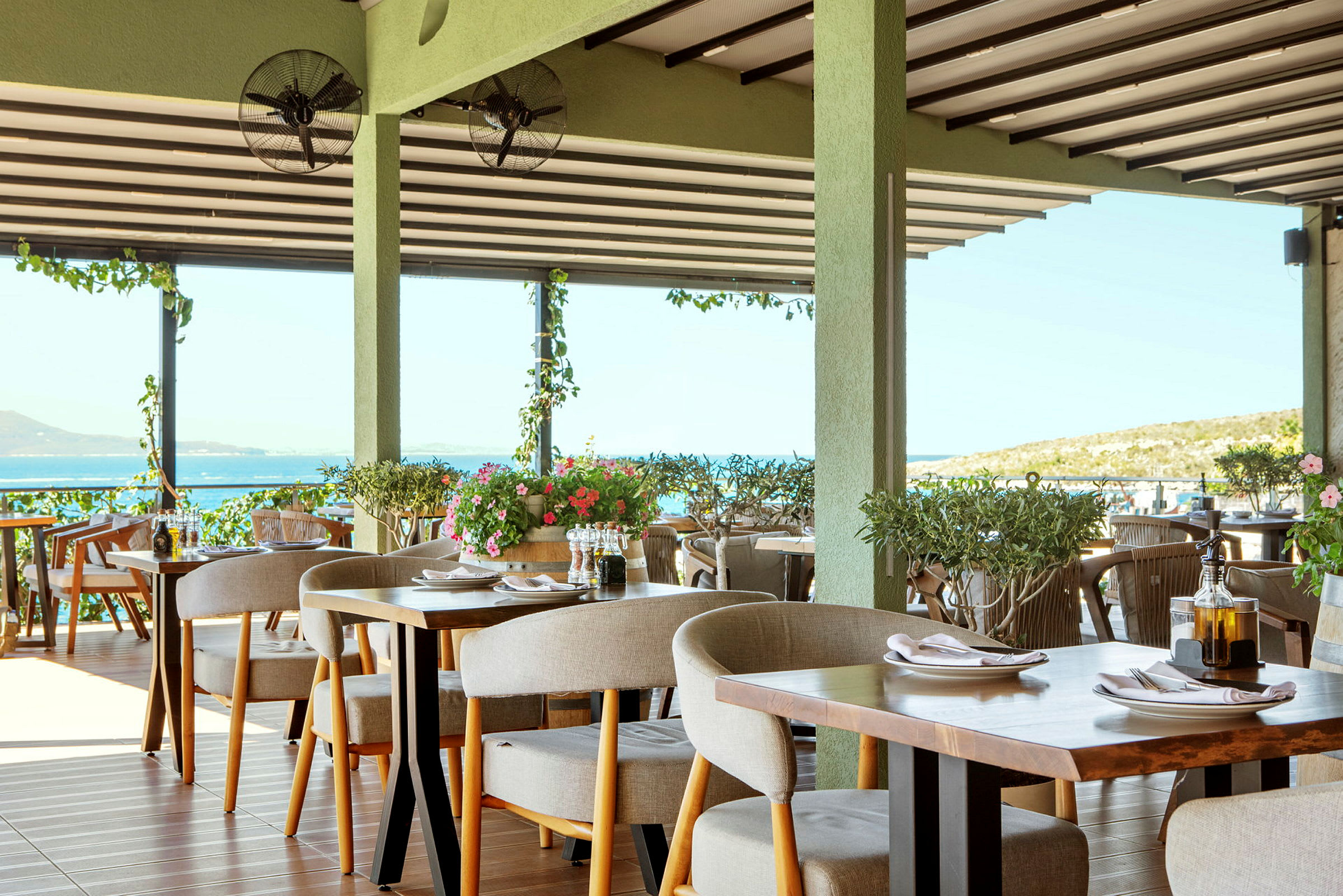 En restaurang med dukade bord och utsikt över havet, dekorerad med gröna växter. (AI-genererad text)