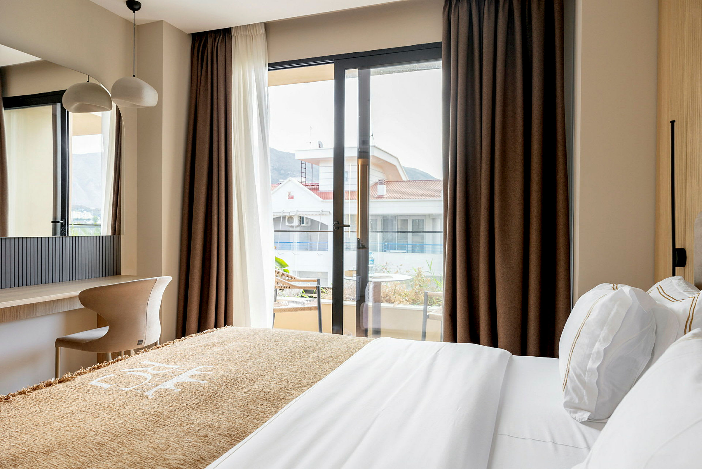 Ett hotellrum med en säng, skrivbord och stol, samt utsikt mot en byggnad genom ett stort fönster. (AI-genererad text)