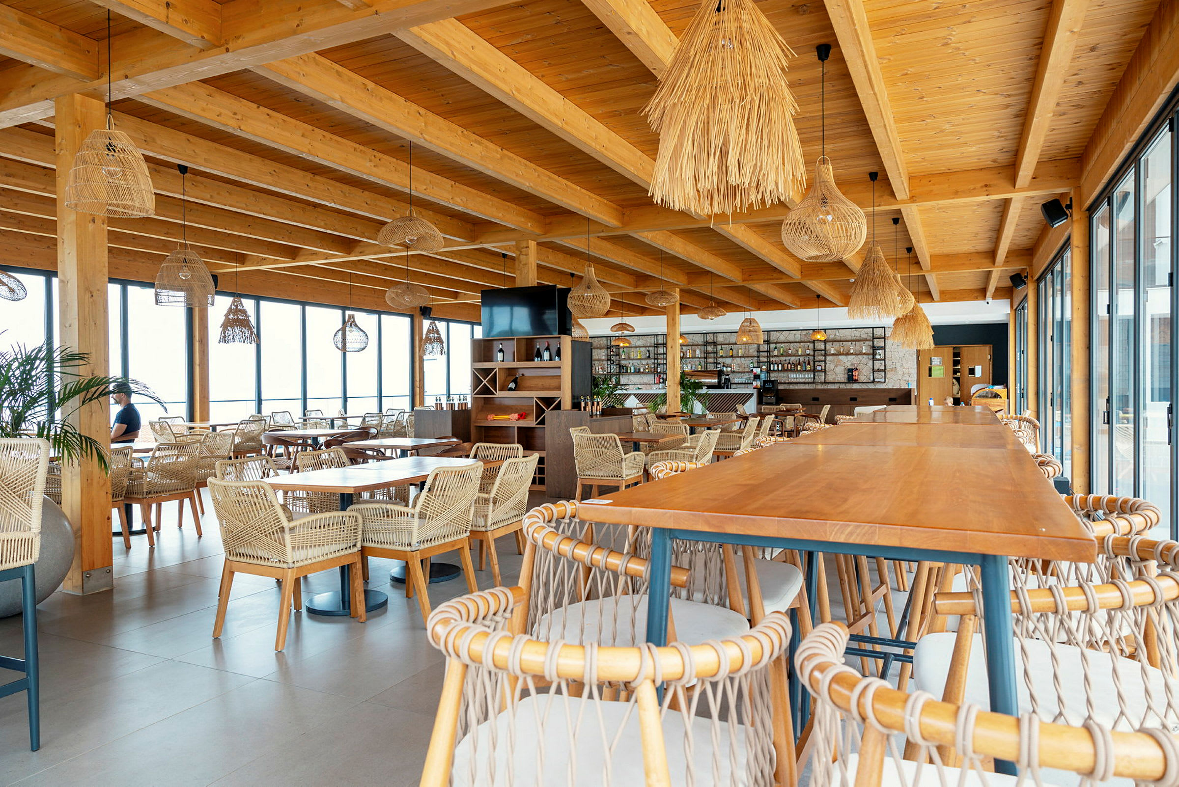 En restaurang med trämöbler och flätade stolar, stora fönster och dekorativa lampor. (AI-genererad text)