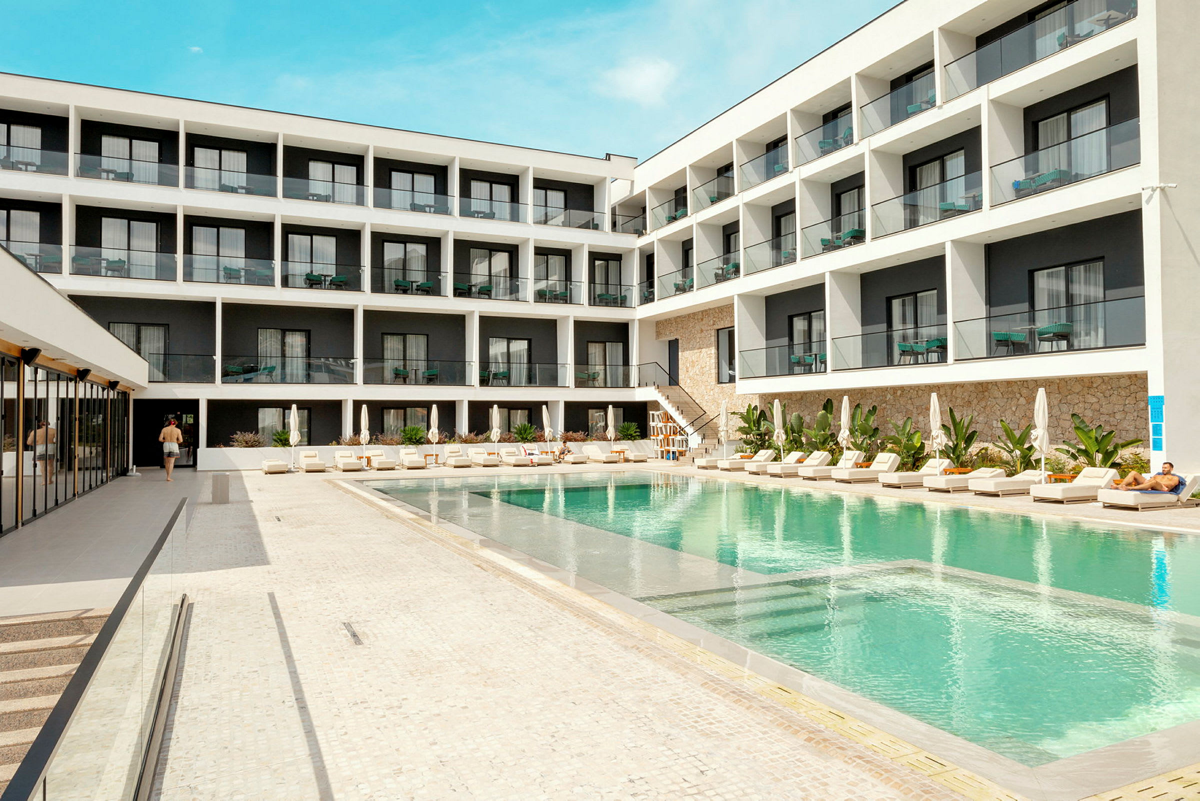 Hotellbyggnad med pool och solstolar i förgrunden. (AI-genererad text)