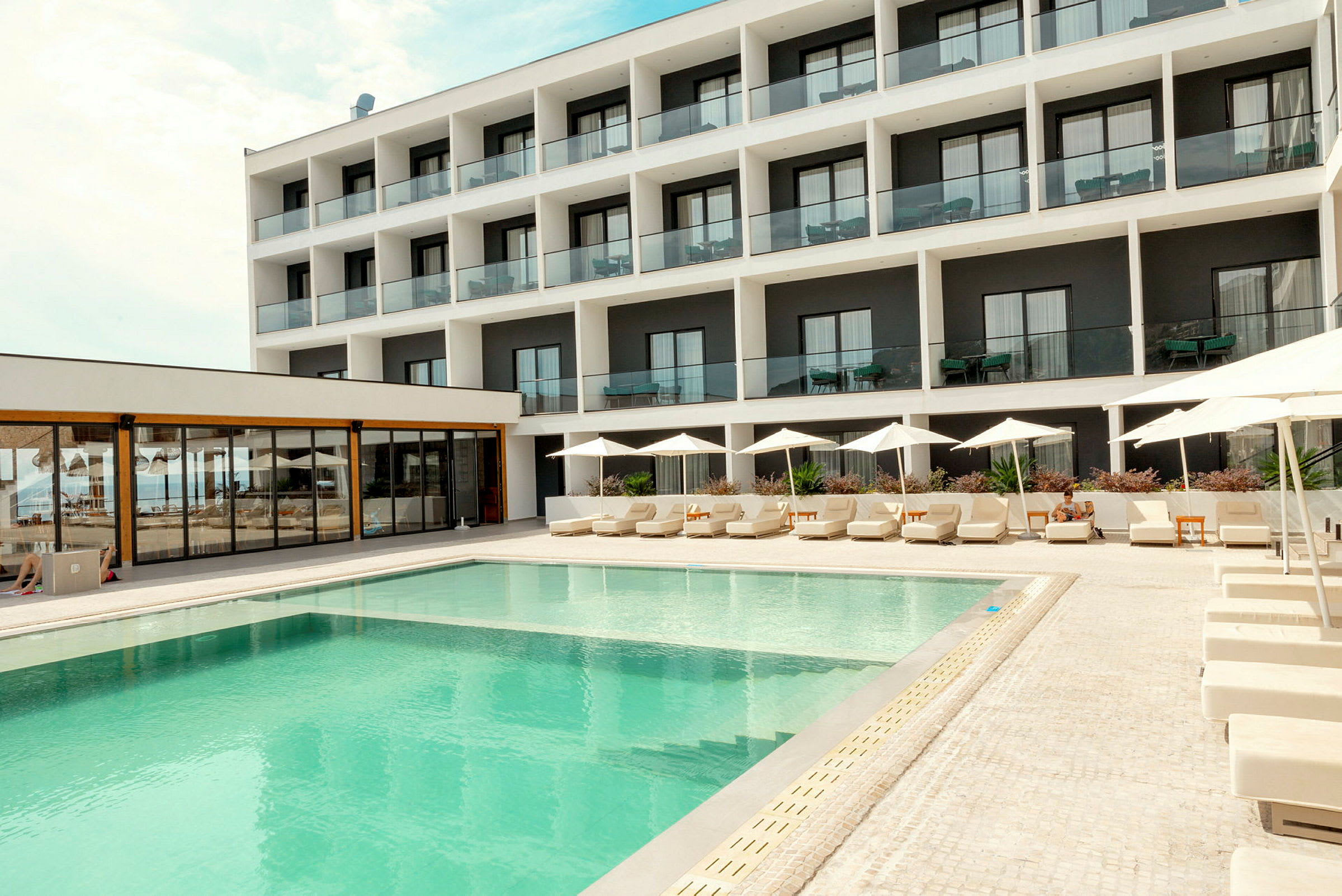 En modern hotellbyggnad med flera våningar, balkonger och en pool framför med solsängar under parasoller. (AI-genererad text)