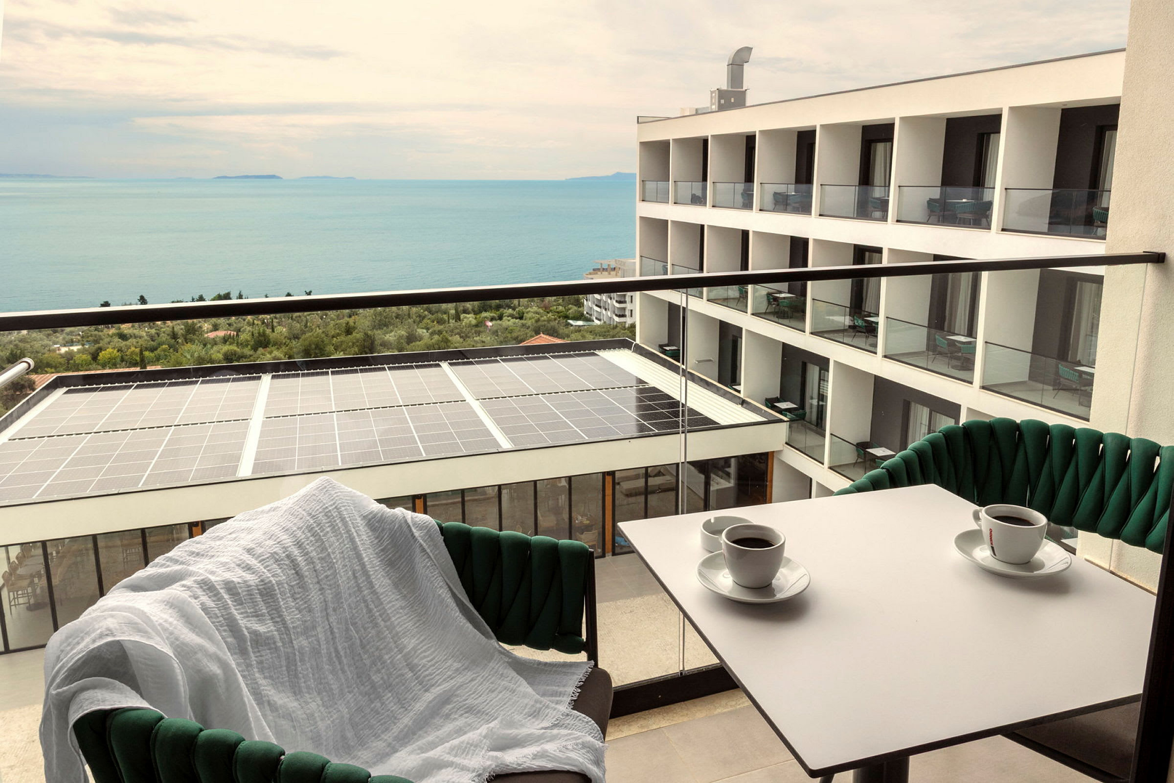 Utsikt från en balkong med två kaffekoppar på bordet, en stol med vit filt, en byggnad och havet i bakgrunden. (AI-genererad text)